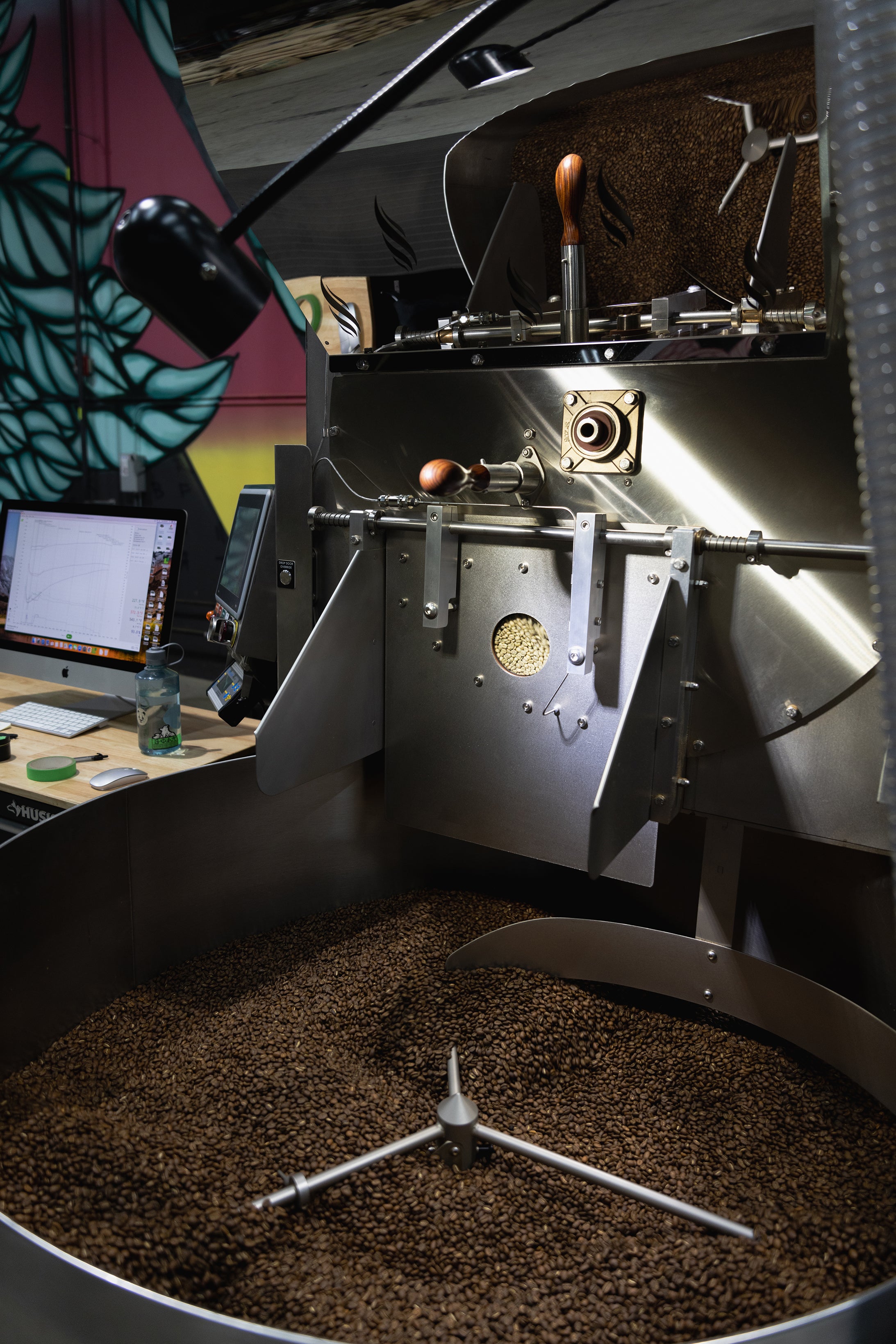 A picture of coffee being roasted in a large roasting tray. There is roasted coffee in the tray and unroasted coffee in the sight glass.