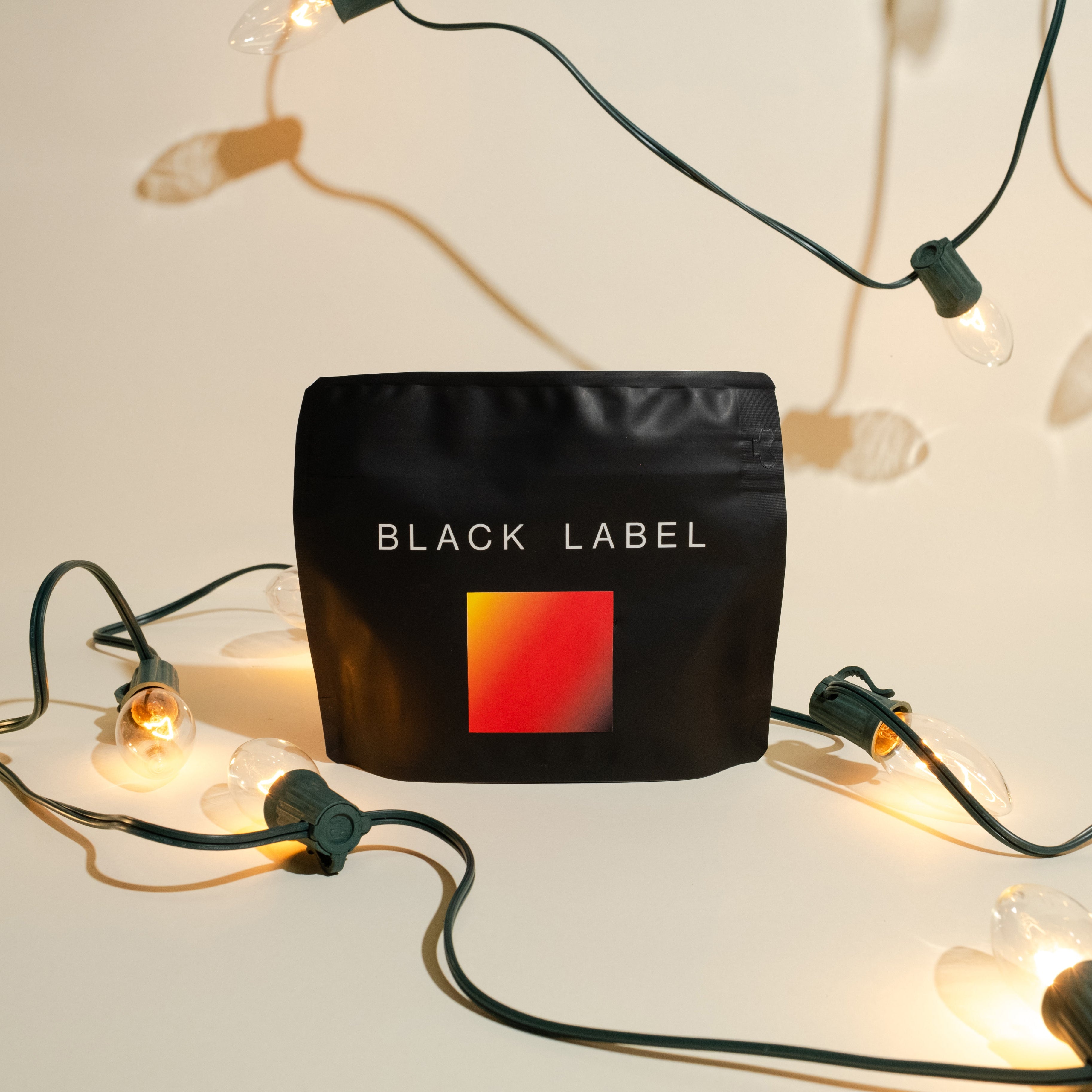 Matte-black pouch labeled “BLACK LABEL” with gradient square, surrounded by warm white lights, featuring Jhonathan Gasca - Natural Pacamara coffee.