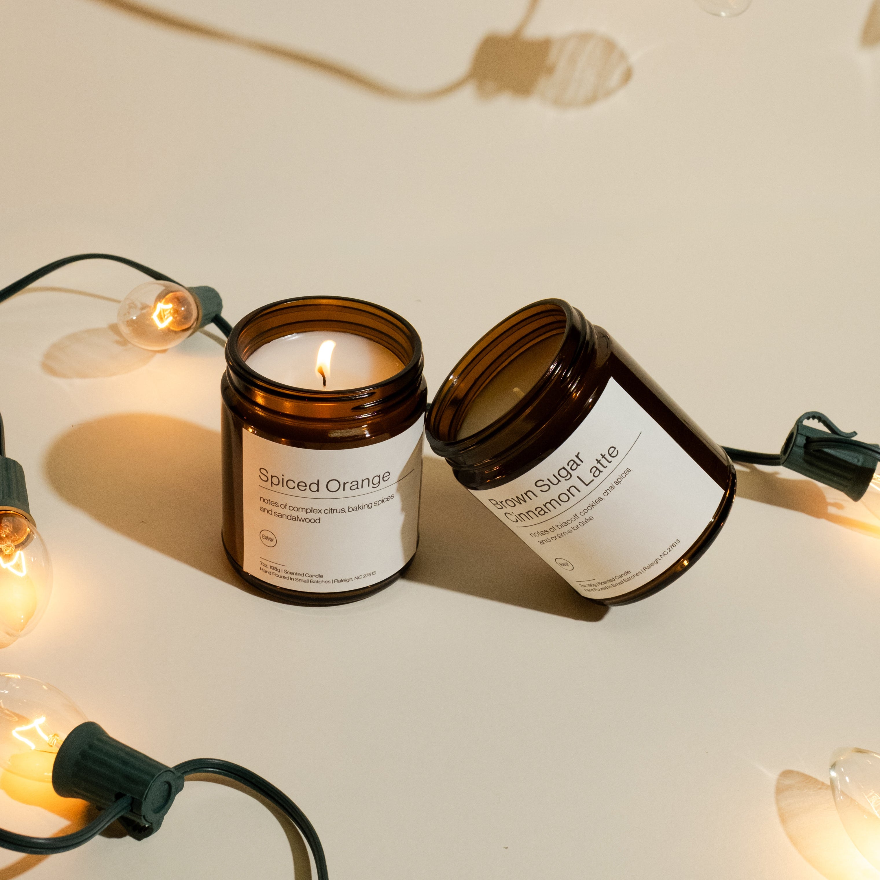 Two candles stacked next to each other on a holiday light themed backdrop. One of the candles is lit and has the label, Spiced Orange.