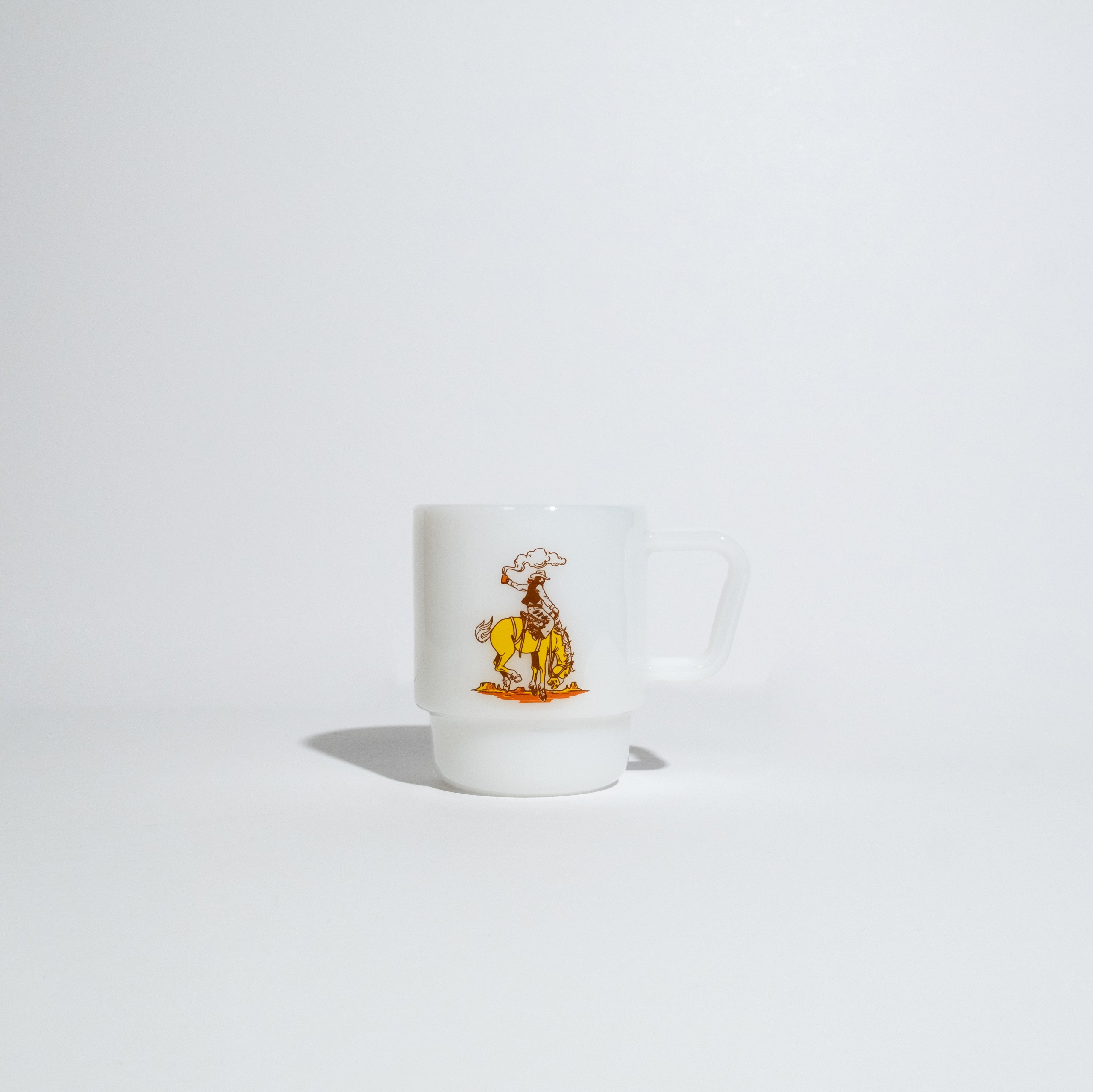 A small white ceramic coffee mug featuring a colorful illustration of a cowboy riding a horse, set against a seamless light gray background.