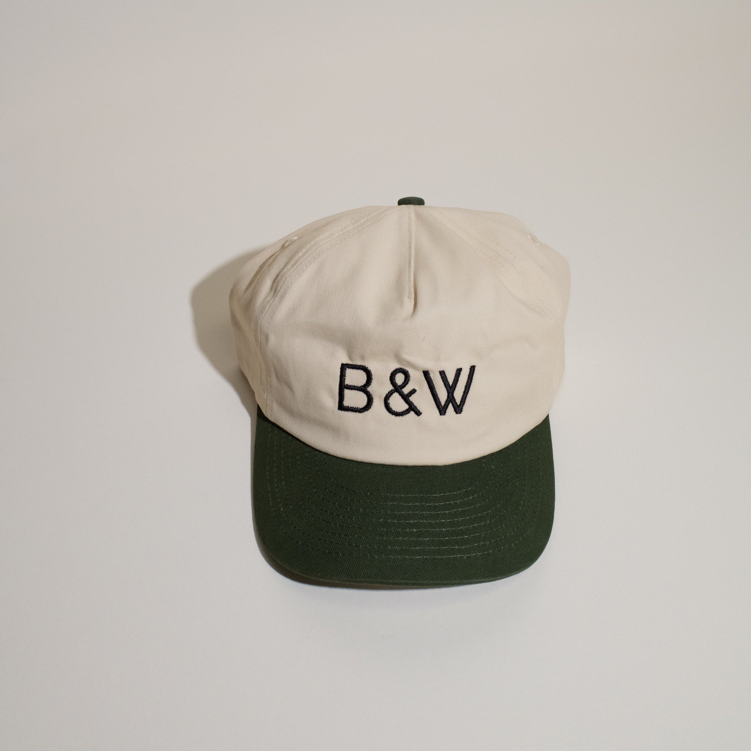 B&W Two Tone Hat with off-white crown, dark green brim, B & W embroidery, adjustable snapback, and mesh liner for structure.