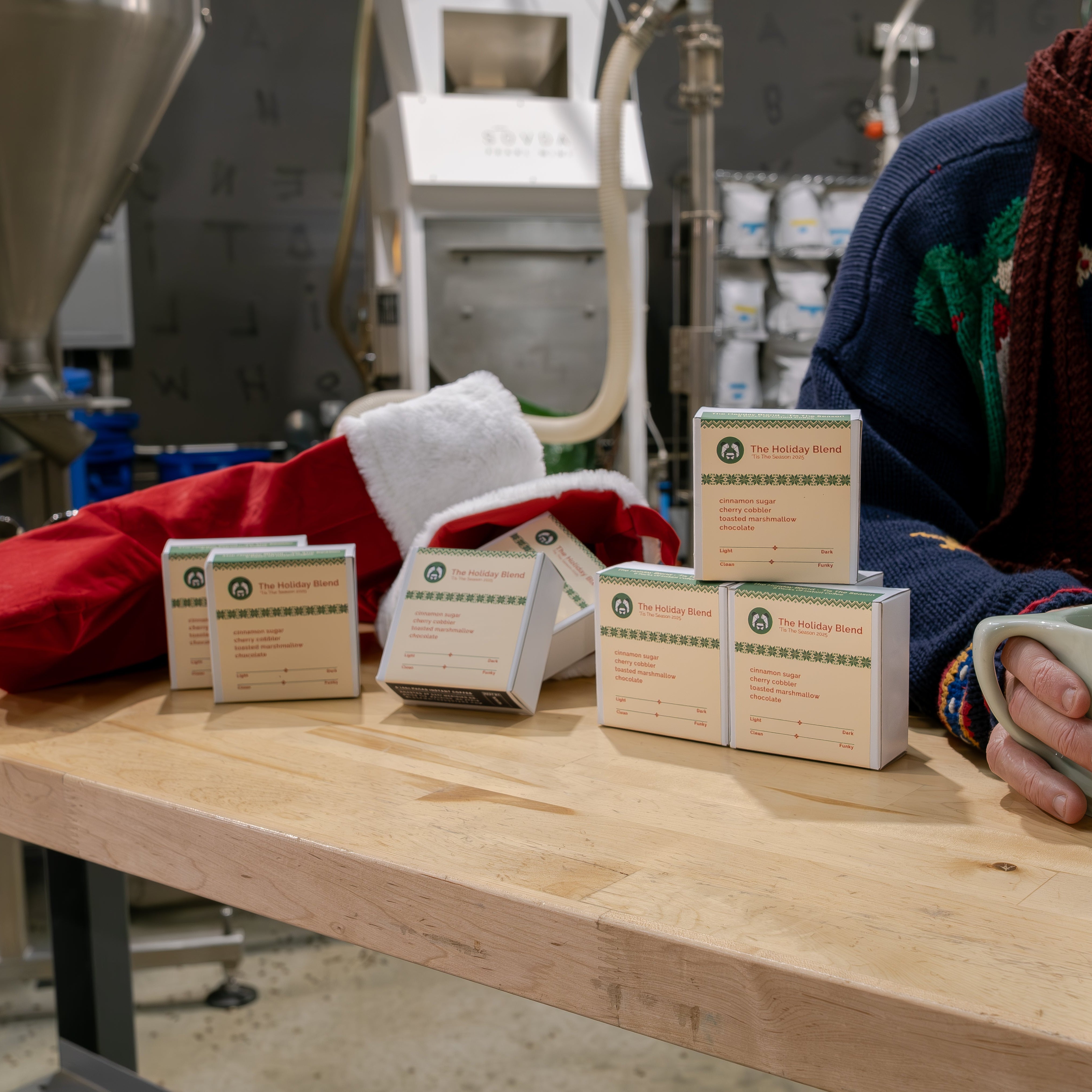 Holiday Blend 'Tis The Season 2025 - Instant Coffee boxes on a countertop with festive stocking; person in sweater holds mug.