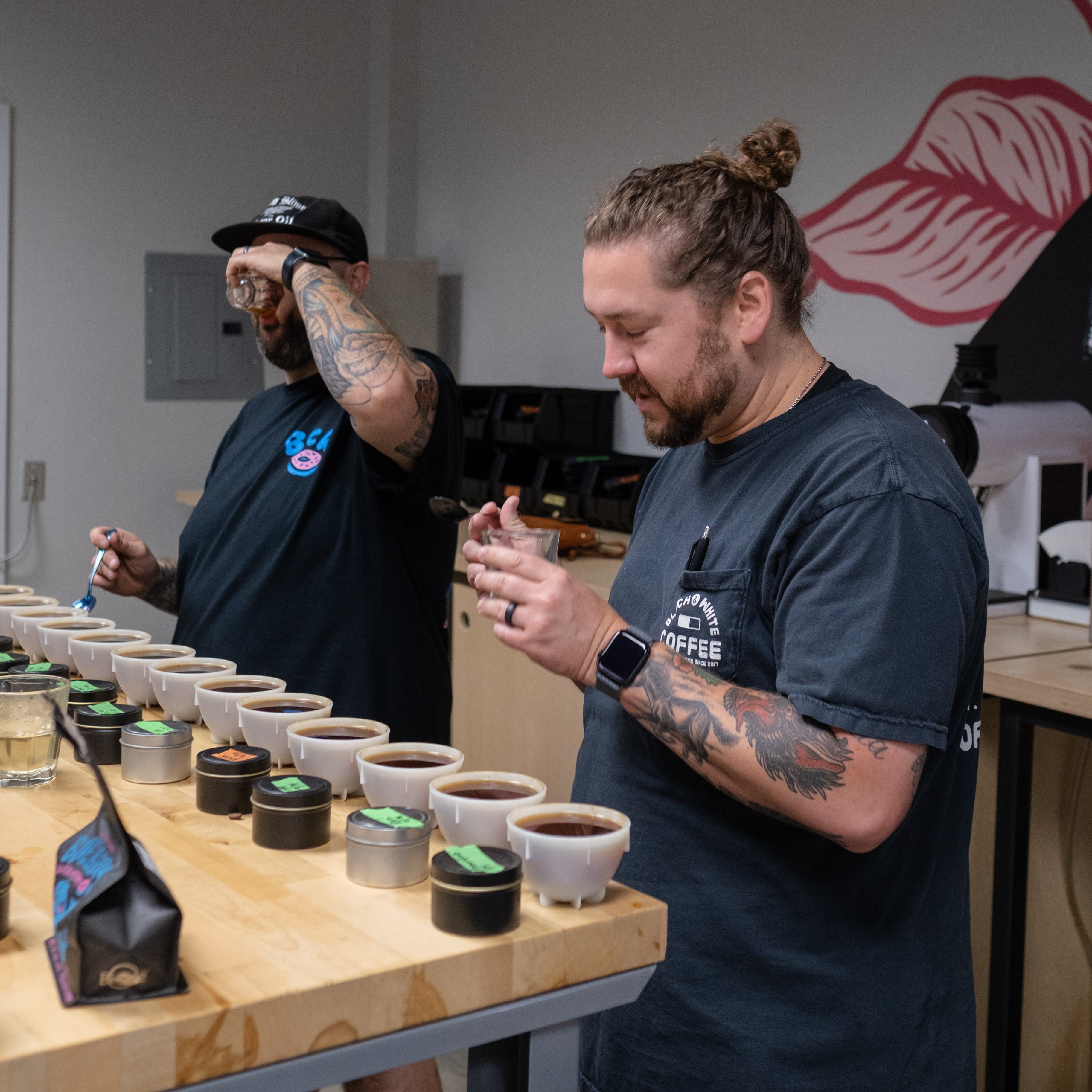 Two men are cupping coffees around a standing table. The table is filled with a selection of different coffees in shallow bowls.