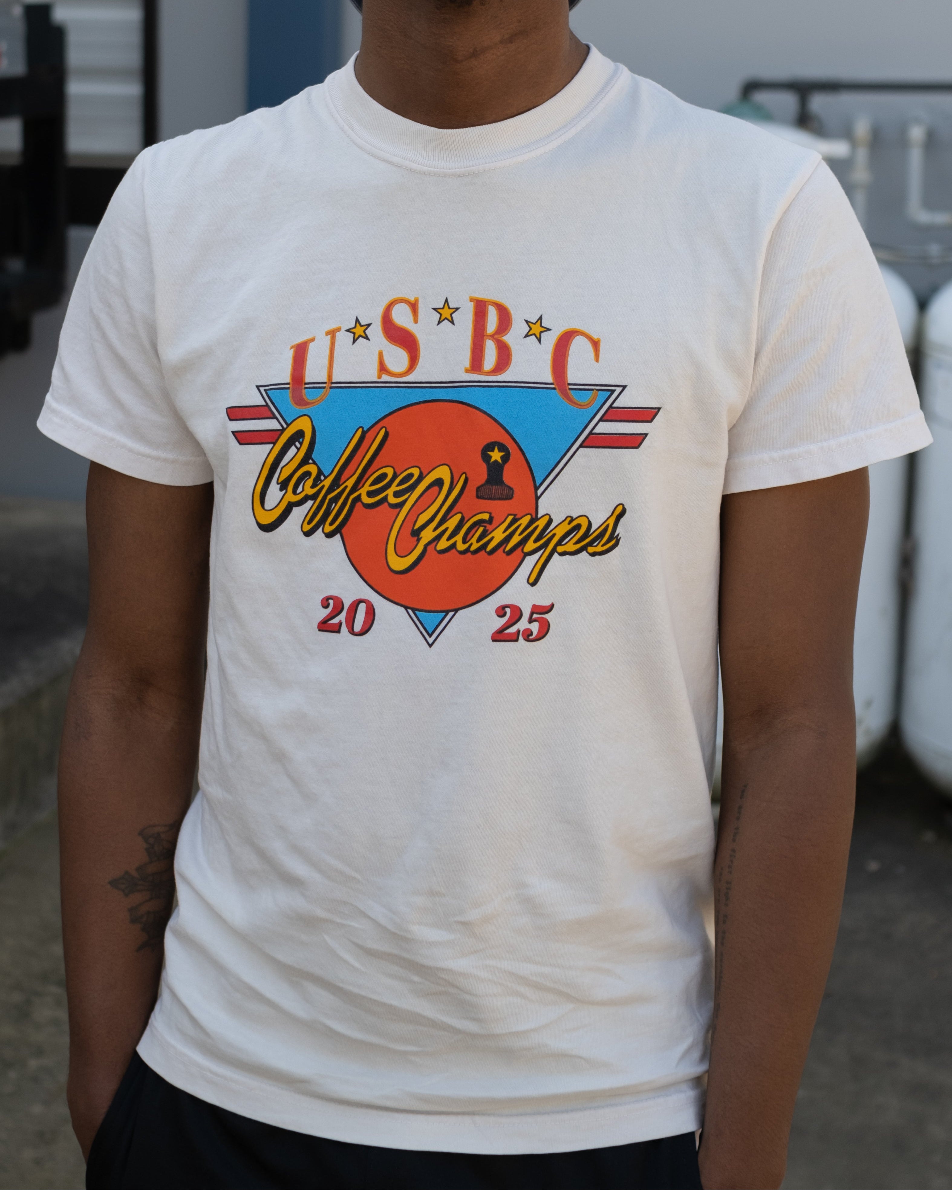 2025 USBC Shirt made of heavyweight, pre-shrunk cotton, features a relaxed fit, twill label, and reinforced neck and shoulders for durability. Worn by a model showcasing the frontside of the shirt with words "U.S.B.C, Coffee Champs, 2025." The graphic features an orange circle over an upside down bright blue triangle with 2 red racing stripe horizontally placed.