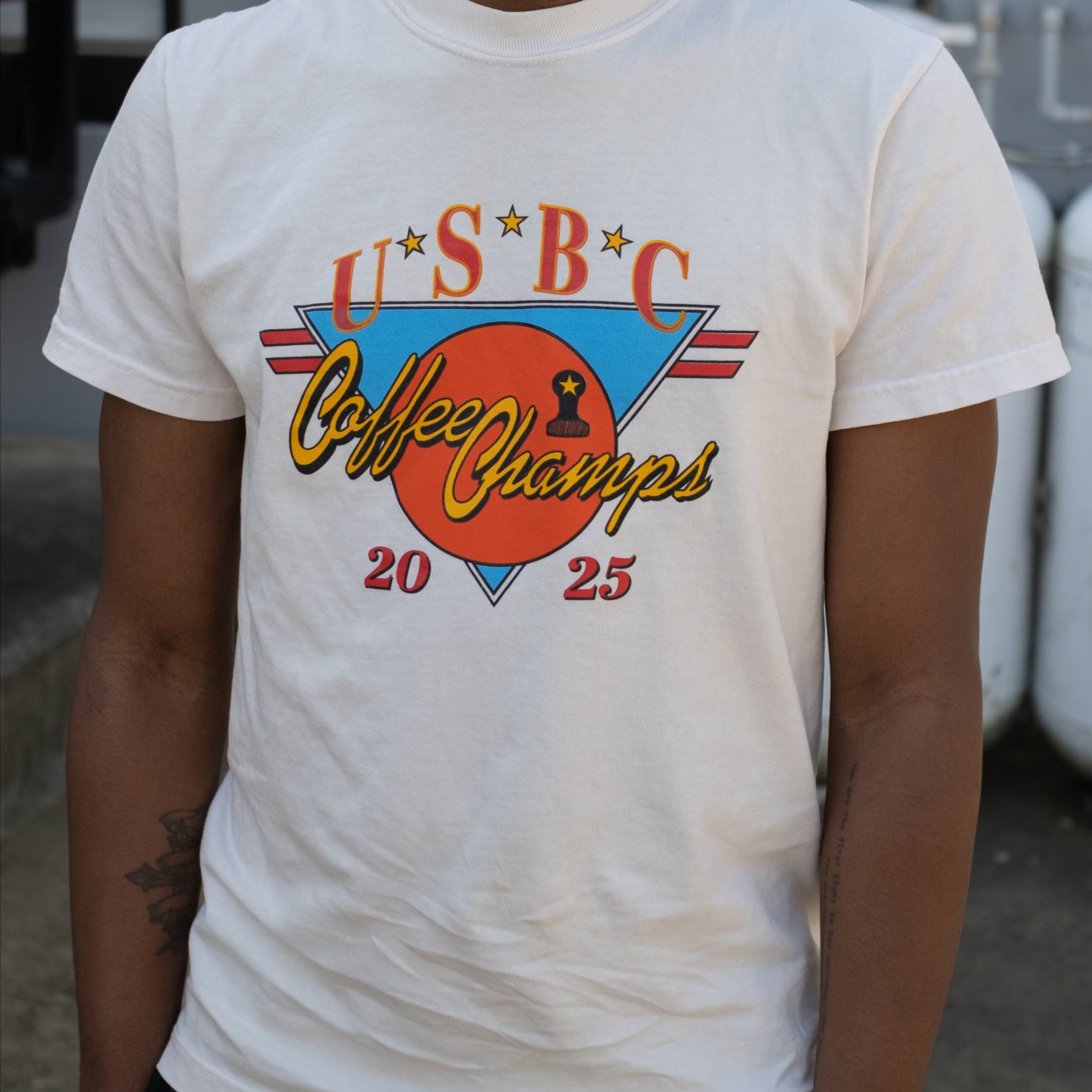 2025 USBC Shirt made of heavyweight, pre-shrunk cotton, features a relaxed fit, twill label, and reinforced neck and shoulders for durability. Worn by a model showcasing the frontside of the shirt with words "U.S.B.C, Coffee Champs, 2025." The graphic features an orange circle over an upside down bright blue triangle with 2 red racing stripe horizontally placed.