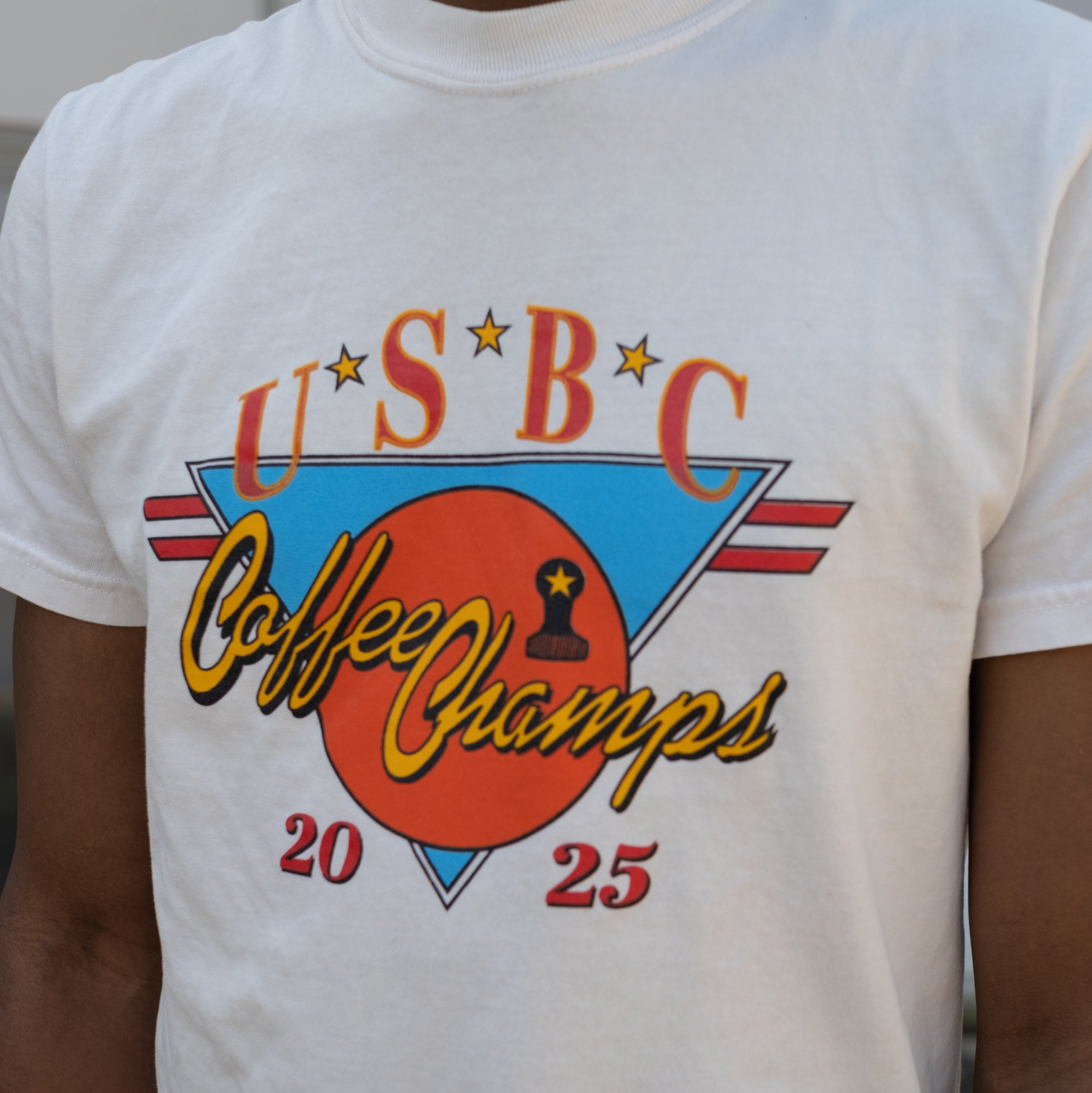 2025 USBC Shirt made of heavyweight, pre-shrunk cotton, features a relaxed fit, twill label, and reinforced neck and shoulders for durability. Worn by a model showcasing the frontside of the shirt with words "U.S.B.C, Coffee Champs, 2025." The graphic features an orange circle over an upside down bright blue triangle with 2 red racing stripe horizontally placed.