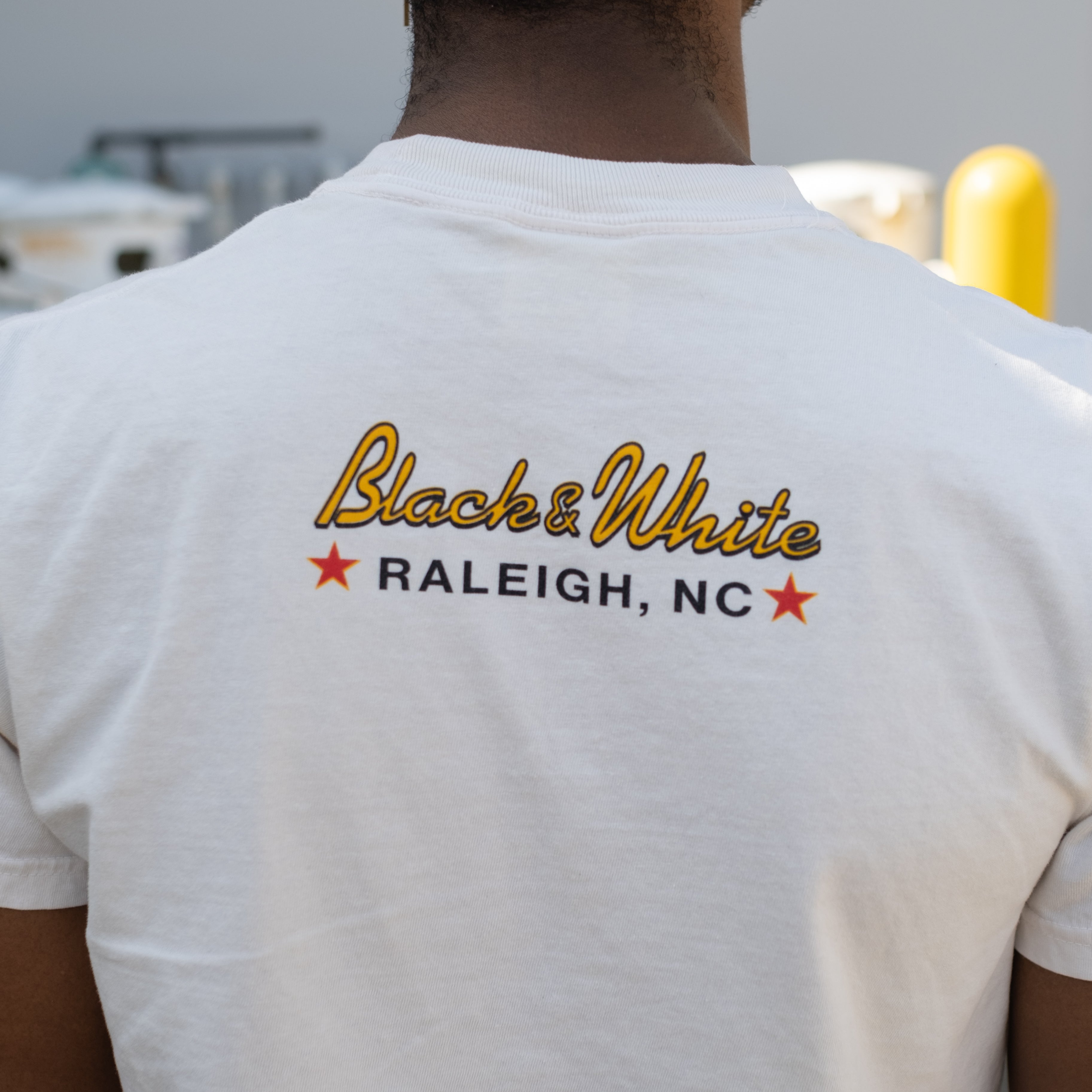 2025 USBC Shirt made of heavyweight, pre-shrunk 100% cotton, featuring a relaxed fit, with a durable twill taped neck and shoulders. Worn by a model showcasing the backside of the shirt with words in script font, "Black & White, Raleigh NC"
