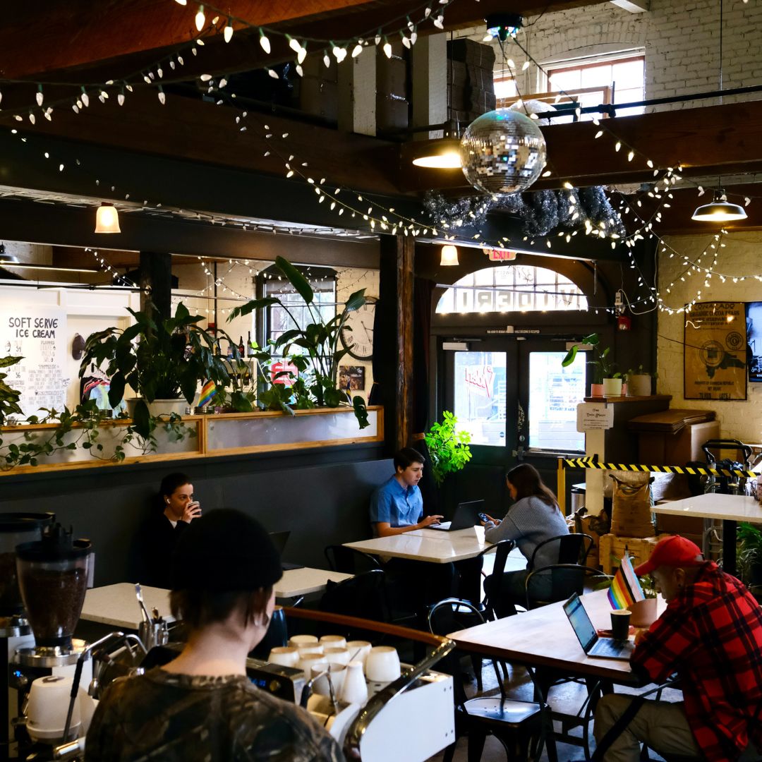 Inside the downtown Raleigh cafe with several tables in the cafe with people sitting at them. The ceiling has market lights and a disco ball hanging in the space. The cafe is darker and has the feel of an older warehouse.