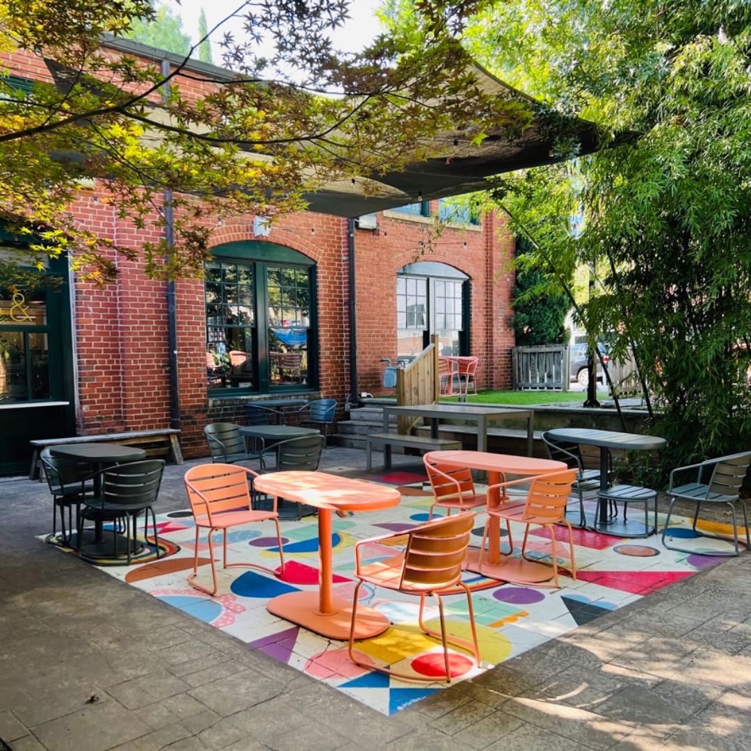Outside courtyard of the downtown Raleigh cafe with a brightly colored geometric design painted on the ground. The chairs are metal and in different colors.