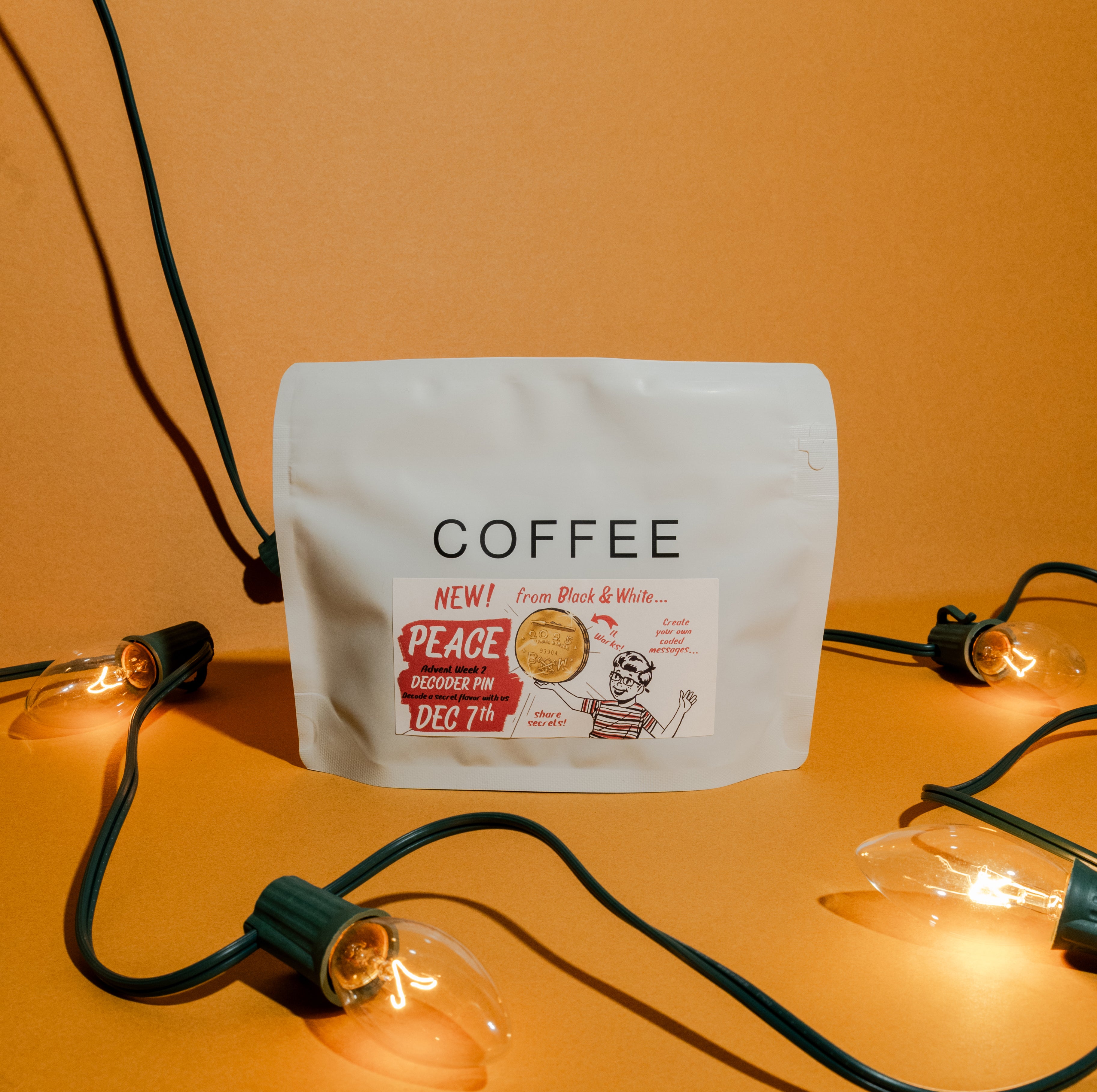 2025 B&W Advent Calendar coffee pouch with “PEACE Advent Week 2 DECODER PIN” sticker, surrounded by decorative light bulbs, against an orange backdrop.