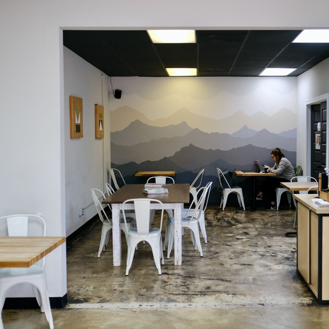 The inside of the Wake Forest cafe with a painted mountain mural in the background in simple colors layered on top of each other. The cafe has white walls and several tables with customers sitting around them.