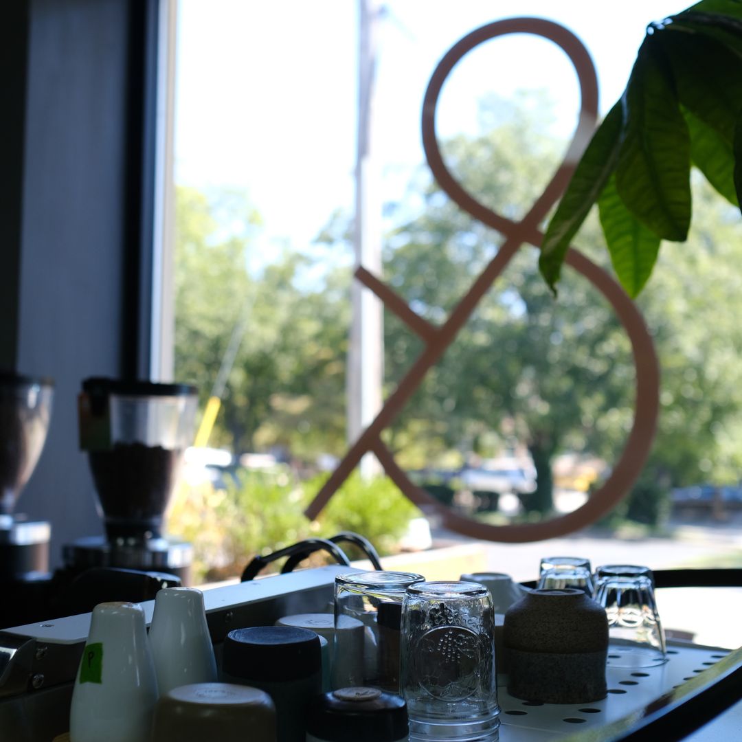 A photo showing the view from inside the cafe, looking out through the large glass window that has an "&" on it in gold. The view includes the top of an espresso machine with ups and class ware on it and the picture of a coffee hopper.