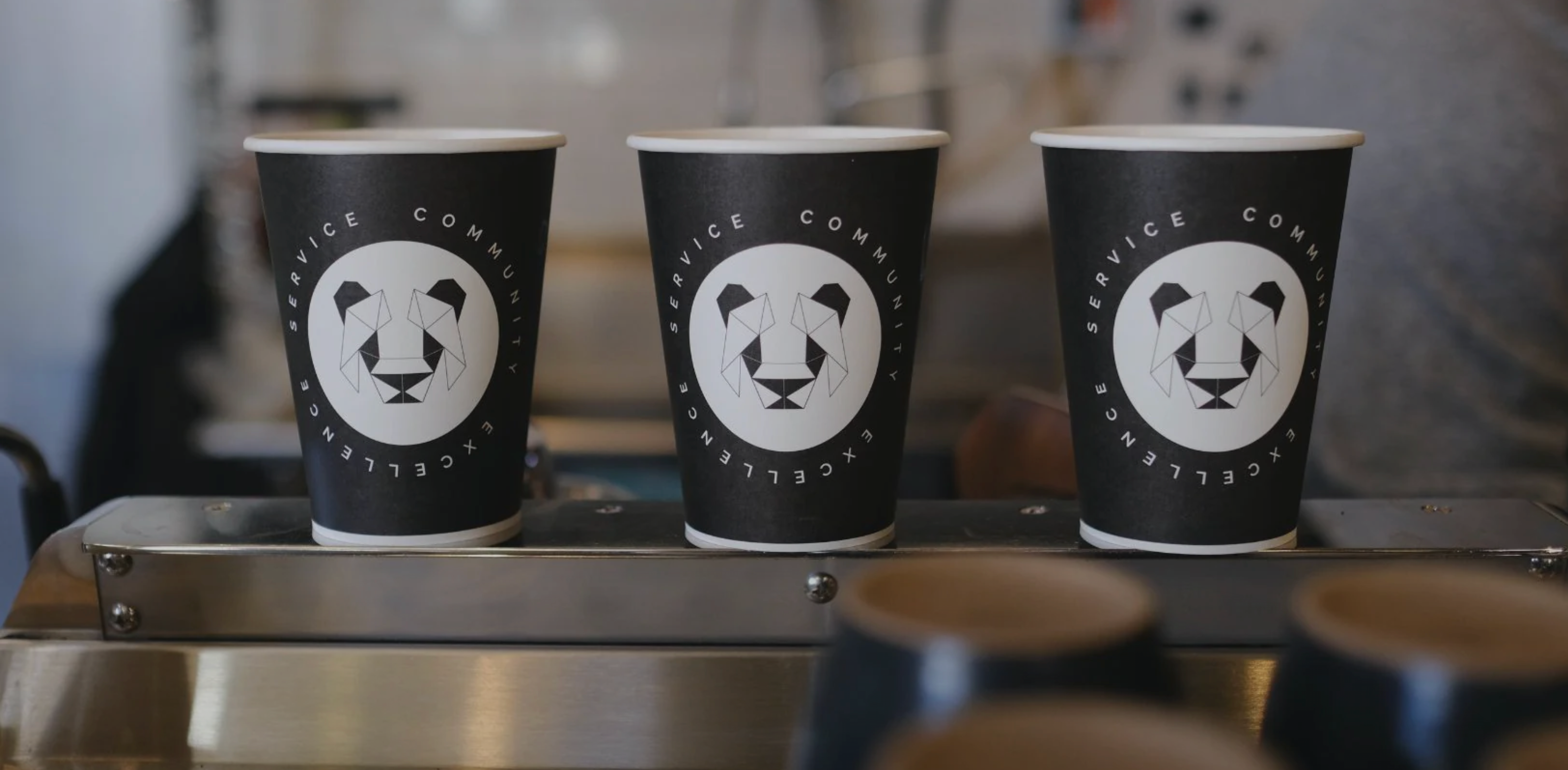 A stack of paper coffee cups with a geometric panda in a white circle with black triangle eyes and black ears. The whole design is symmetrical  and made of various triangles. Around the white circle are the words Service, community and excellence.