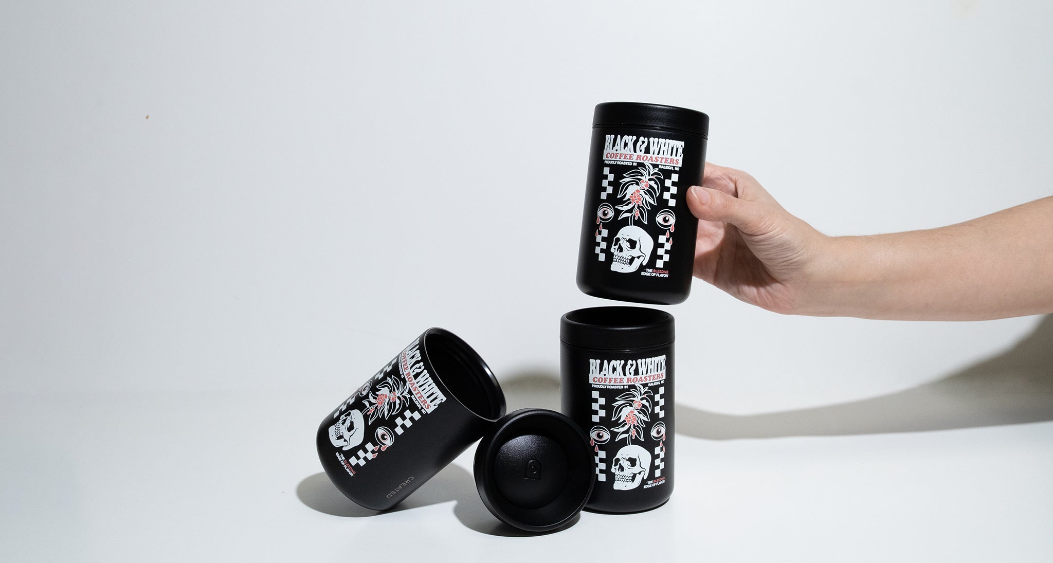 Hand holding a small black Tumbler that says Black & White Coffee Roasters, Bleeding edge of flavor. The hand is stacking the Tumbler on other tumblers.