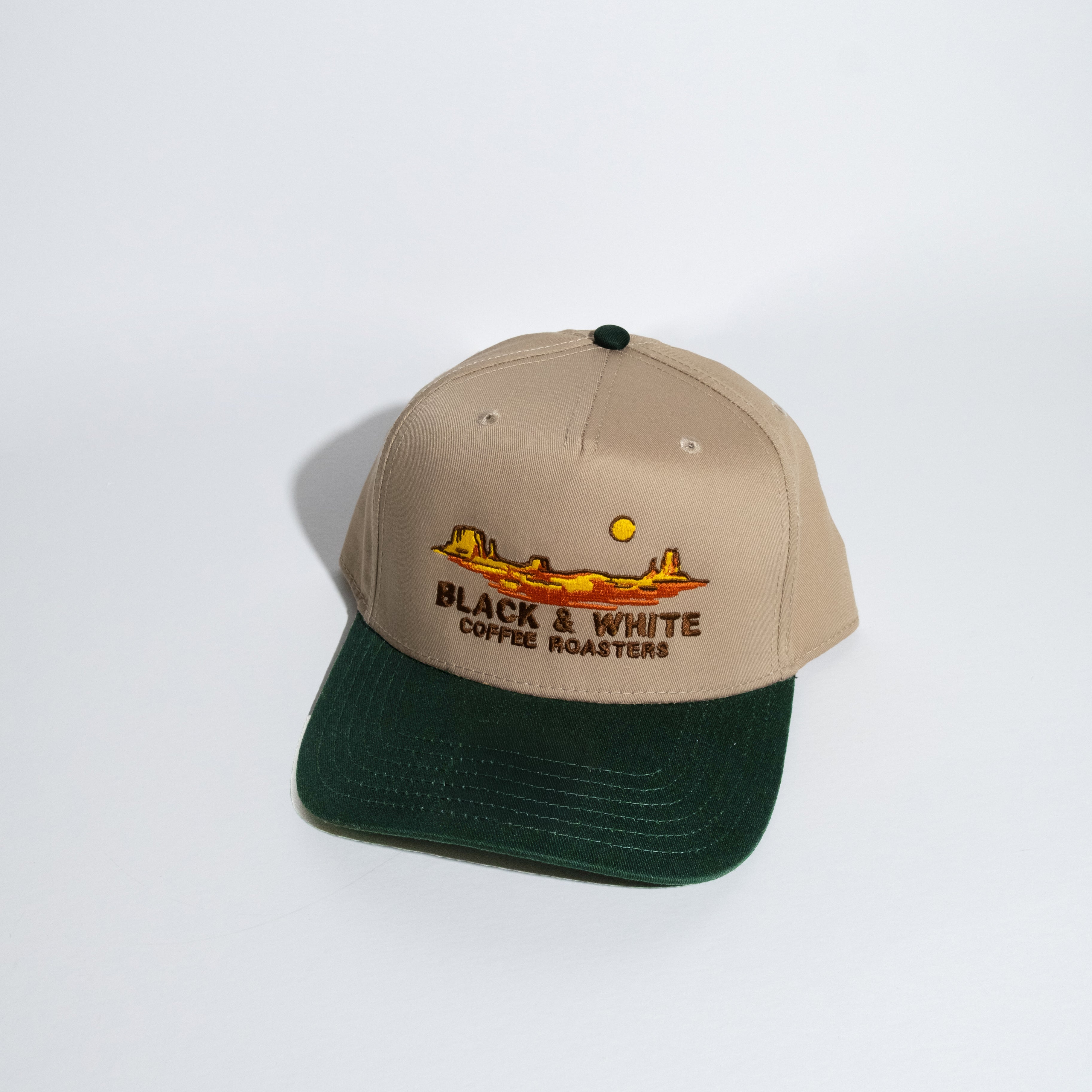 A structured baseball cap with a beige crown and a dark forest green curved brim. The front of the cap features an embroidered graphic of a desert landscape with a yellow sun and the text 'BLACK & WHITE COFFEE ROASTERS'.