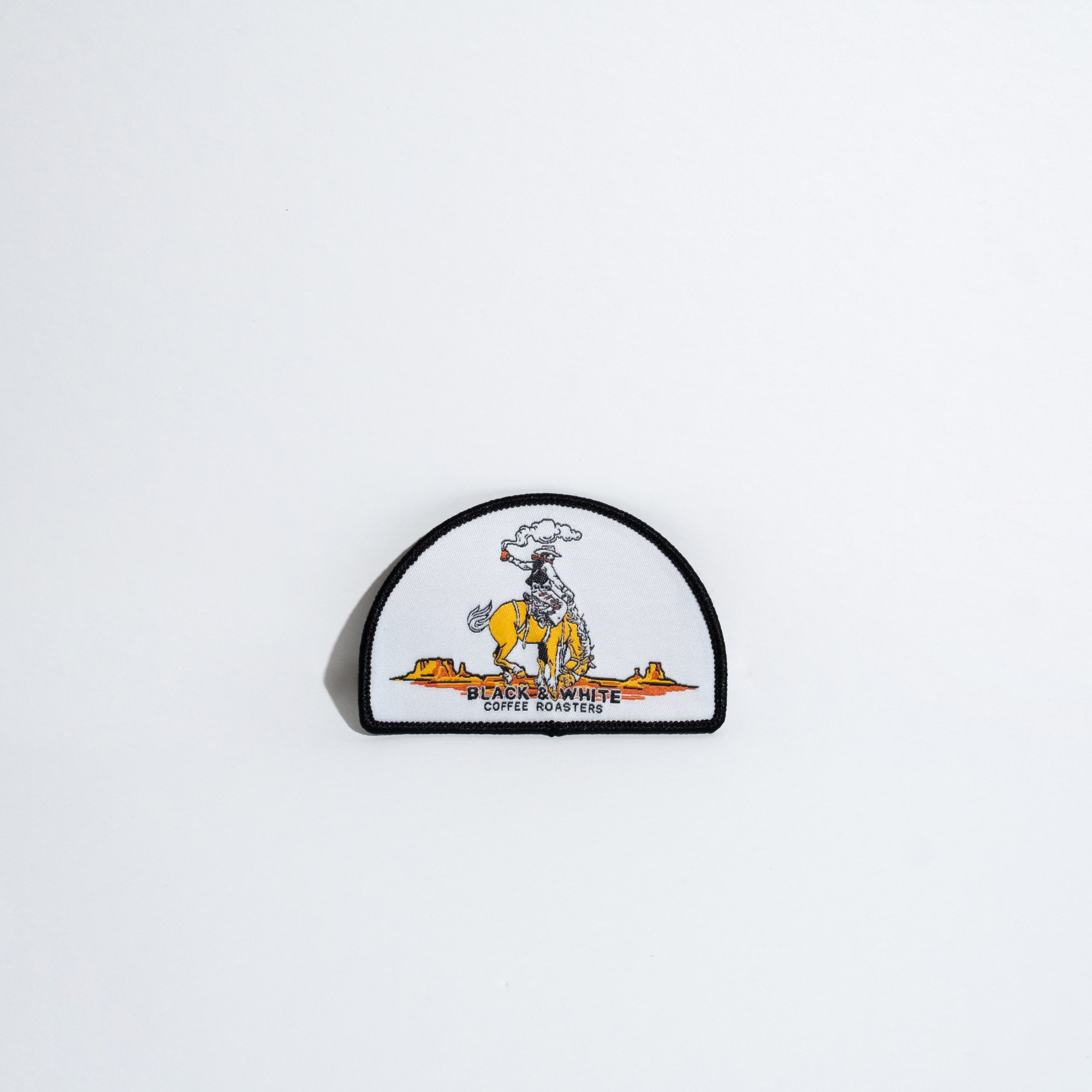 A small embroidered patch featuring a cowboy riding a yellow bucking horse, with the text 'BLACK & WHITE COFFEE ROASTERS' below, shown against a plain white background.