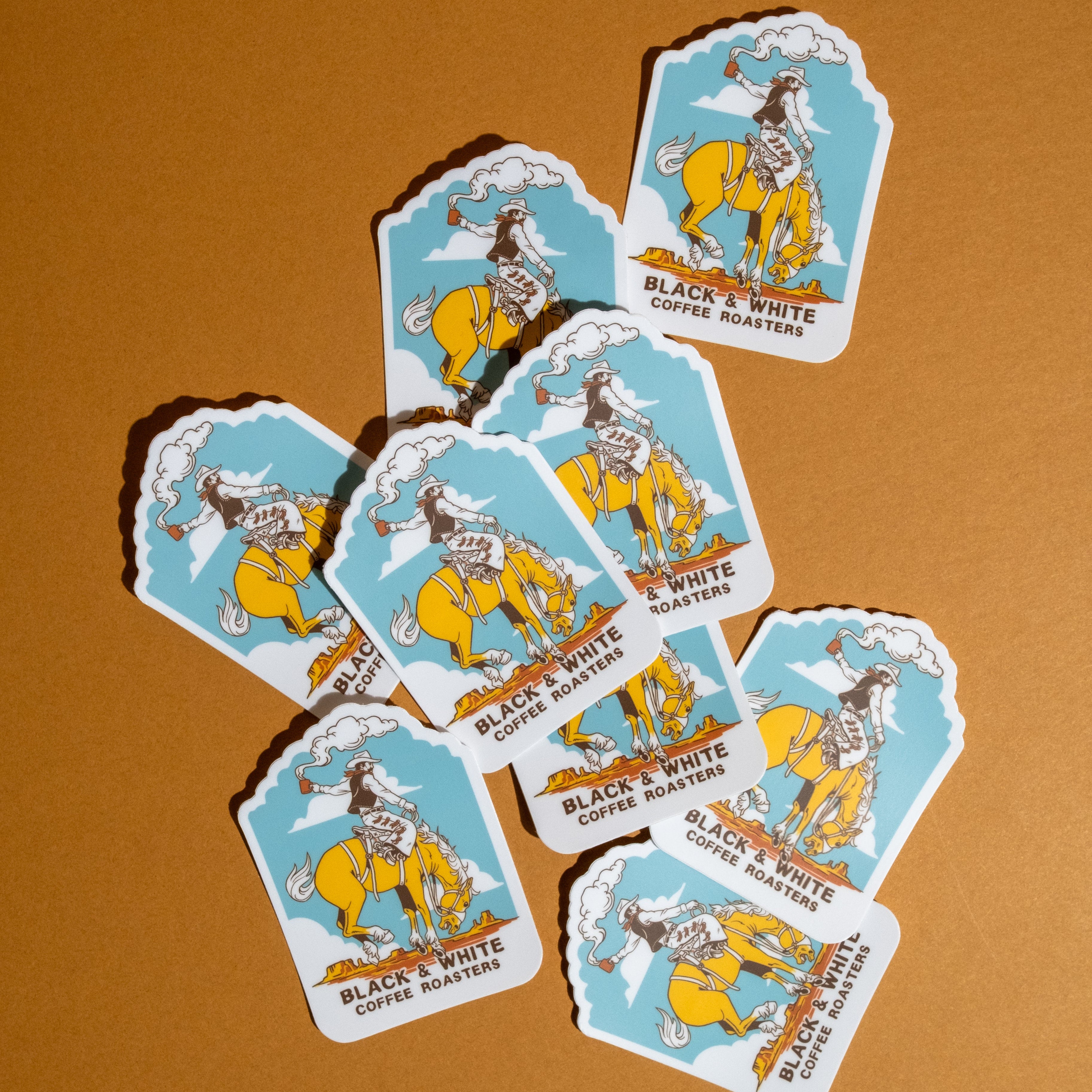 Nine vintage-style die-cut stickers featuring a cowboy riding a yellow horse on a light blue background, with the text 'BLACK & WHITE COFFEE ROASTERS,' arranged on a warm brown surface.