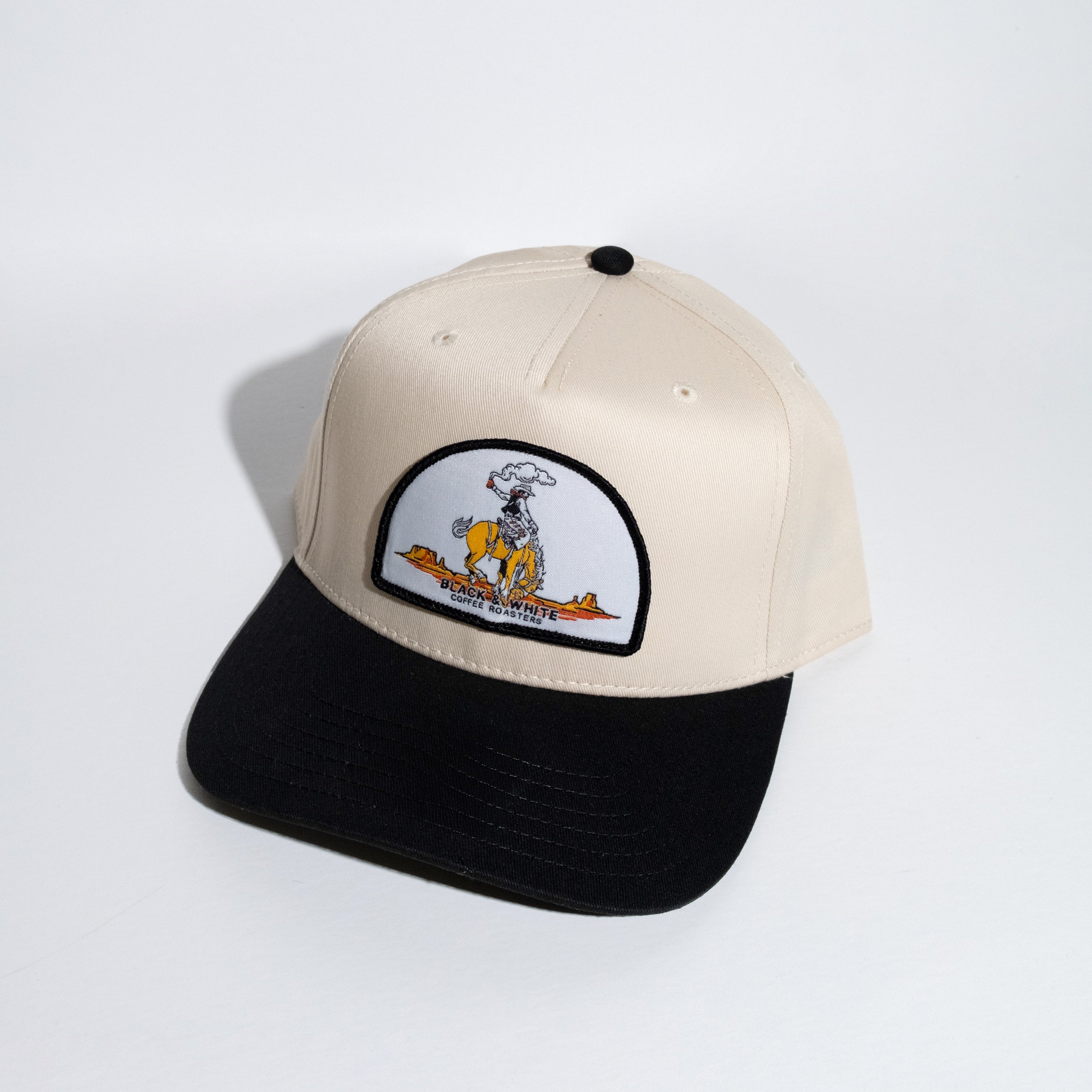 A structured baseball cap with a cream-colored crown and a black flat brim, featuring a circular embroidered patch on the front with a cowboy cartoon character.