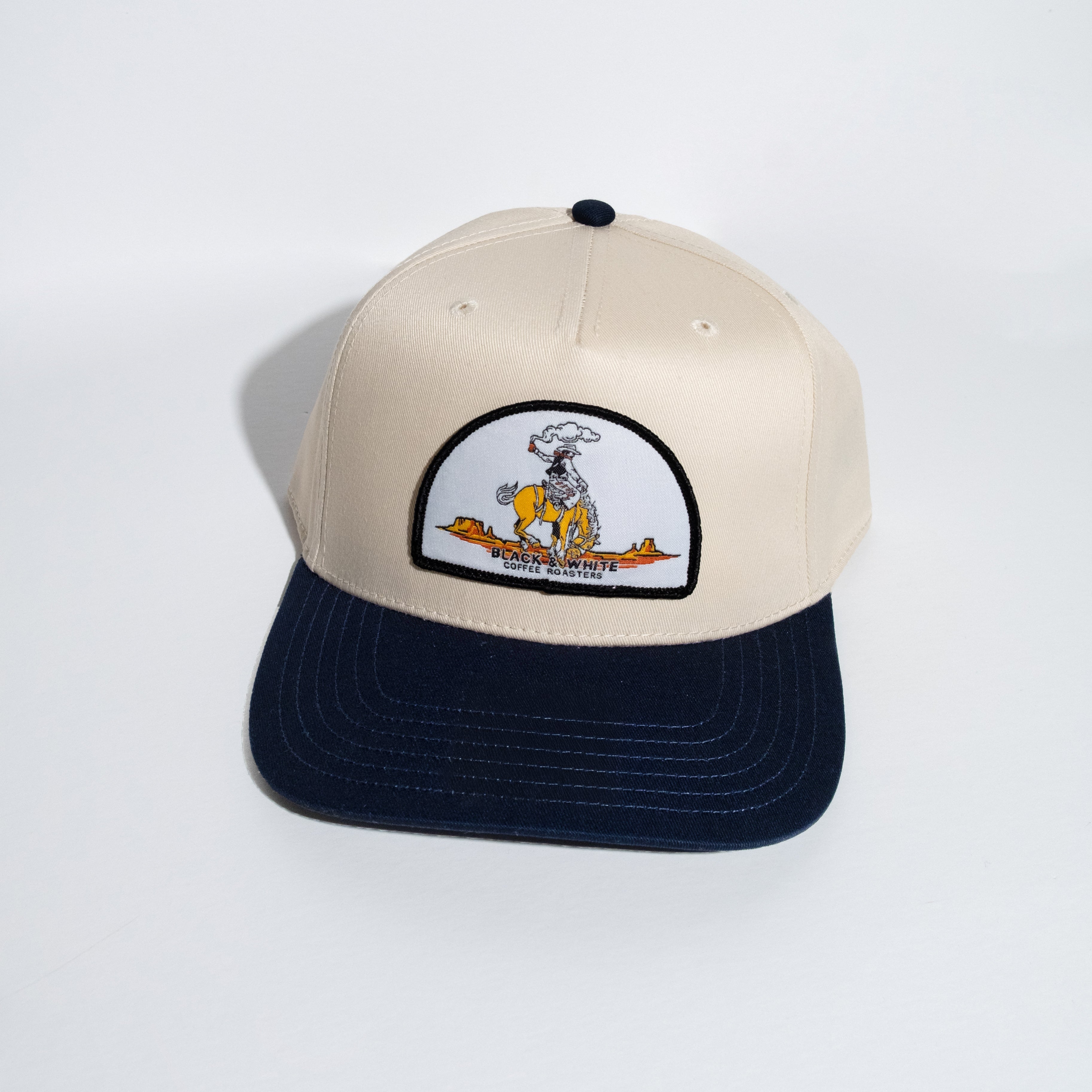 A structured snapback cap with a cream-colored crown and a dark navy blue flat brim. The front of the crown features a white arched patch with a cowboy riding a horse.