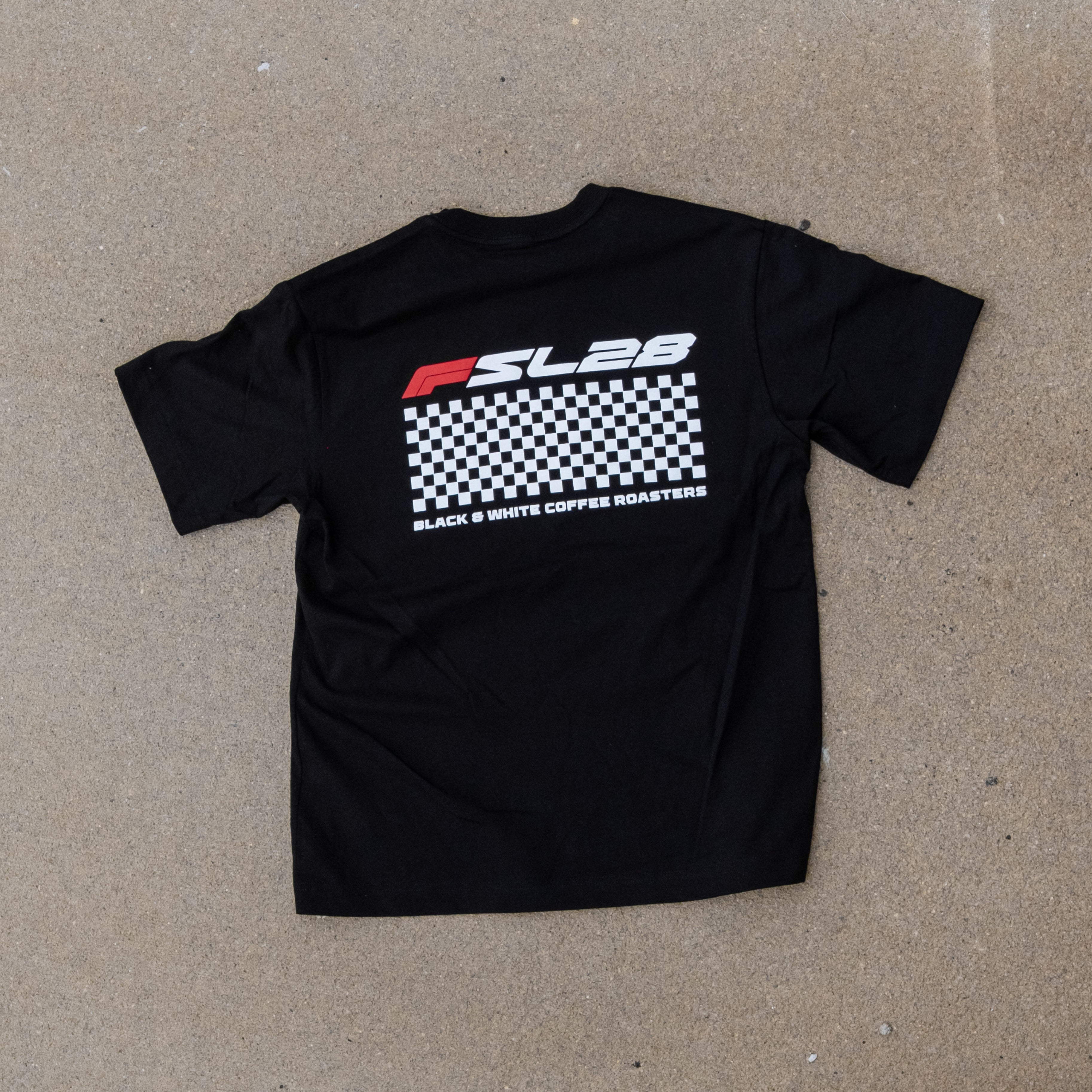 FSL28 Birthday Shirt featuring a red and white logo with BLACK & WHITE COFFEE ROASTERS above a checkerboard graphic on the back, laid on concrete.