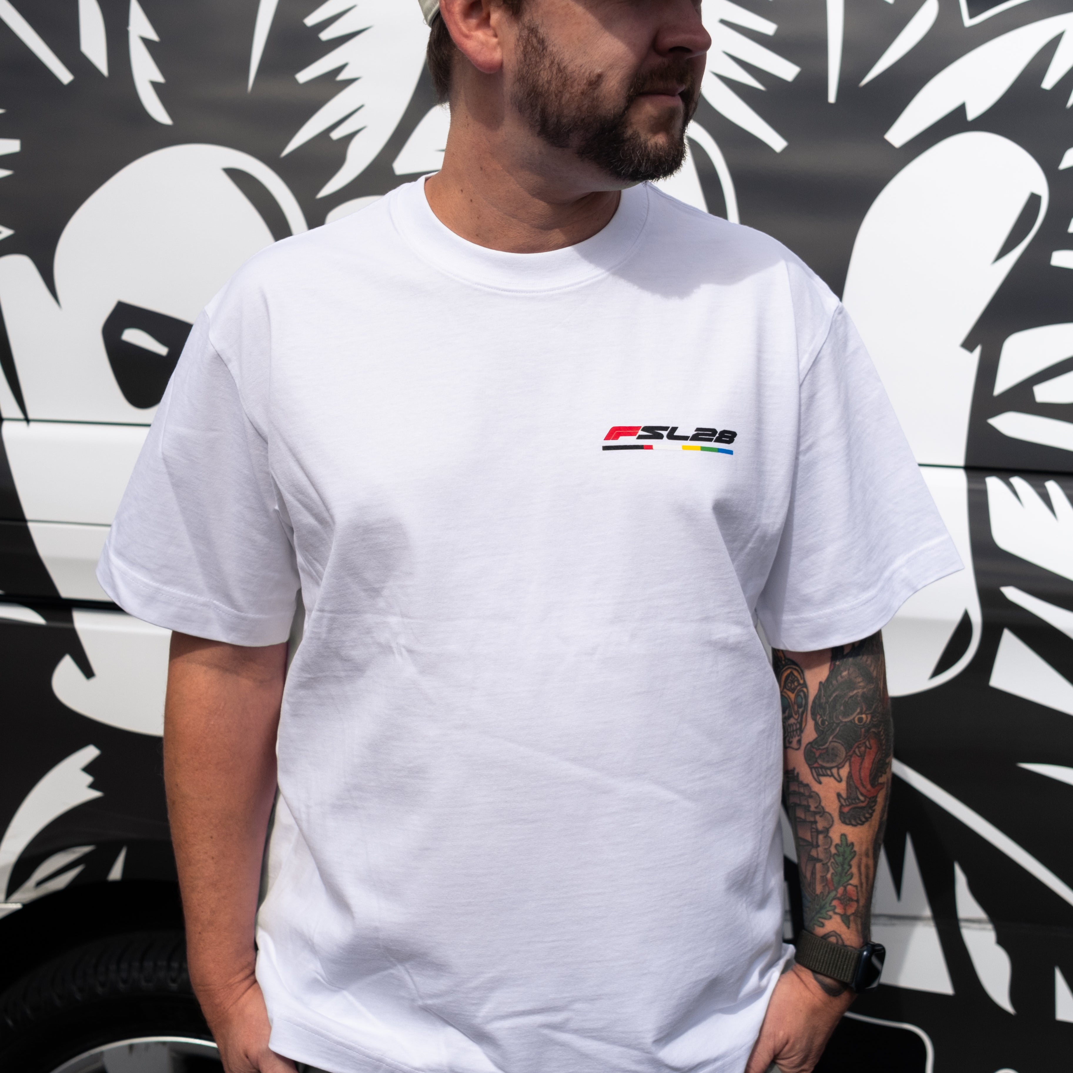 Man wearing Color Bar Birthday Shirt with FSL28 logo, showcasing tattooed forearm and smartwatch, stands against a bold graphic backdrop.