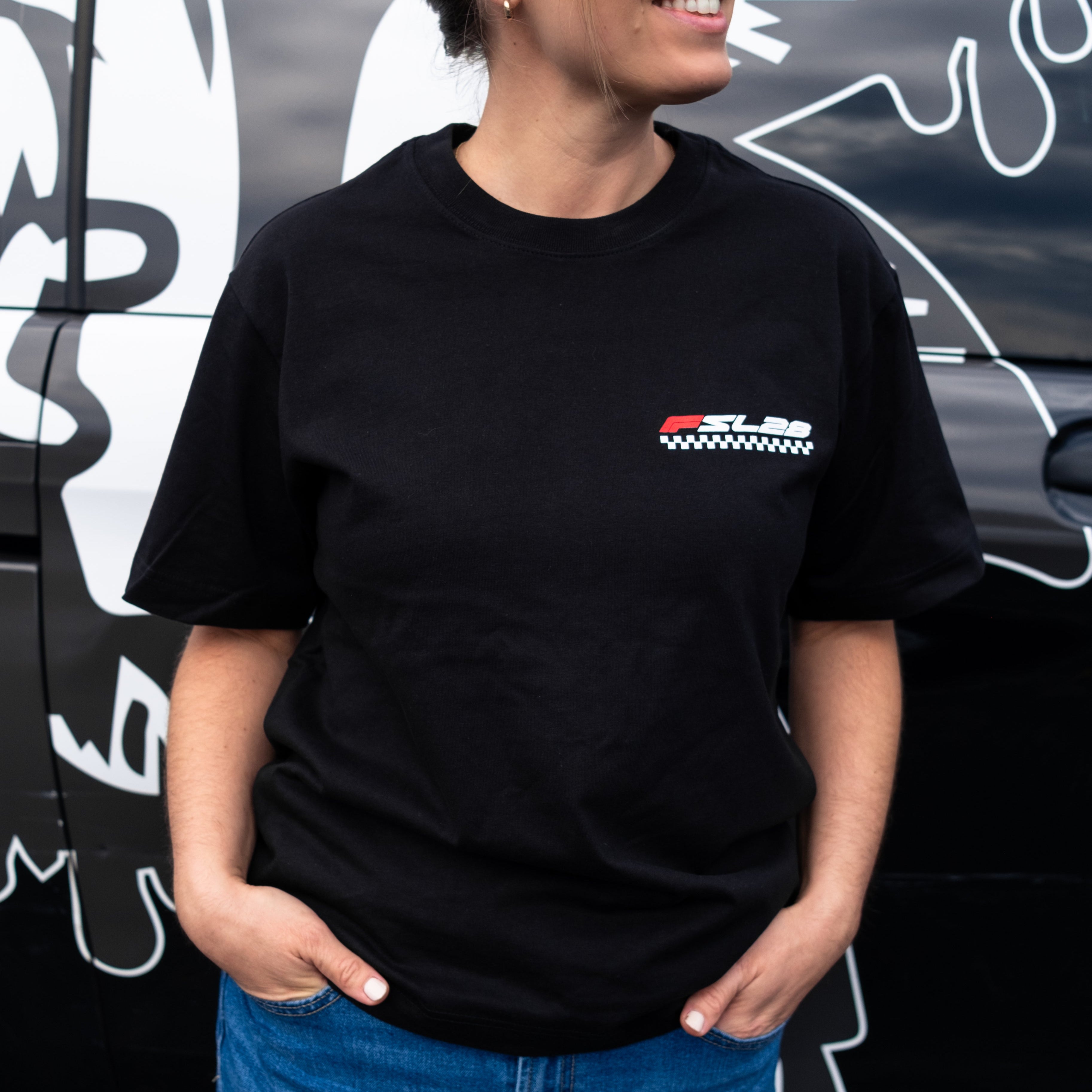 Person wearing the FSL28 Birthday Shirt, featuring a SL28 logo over a checker-stripe, paired with blue jeans, standing before a graphic-adorned vehicle.
