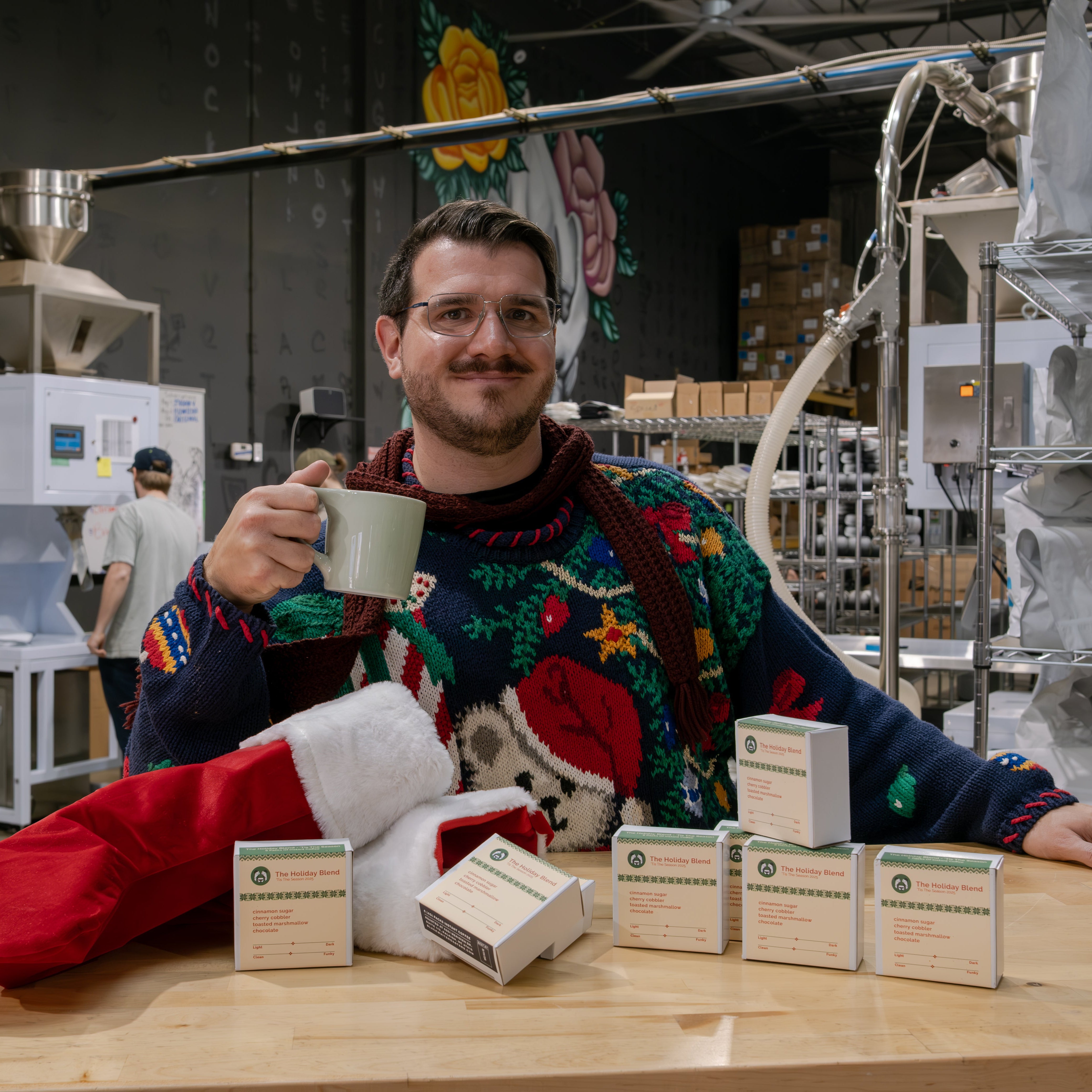 Man toasting with mug behind worktable, showcasing Holiday Blend 'Tis The Season 2025 instant coffee boxes and Santa hat, in a production area.
