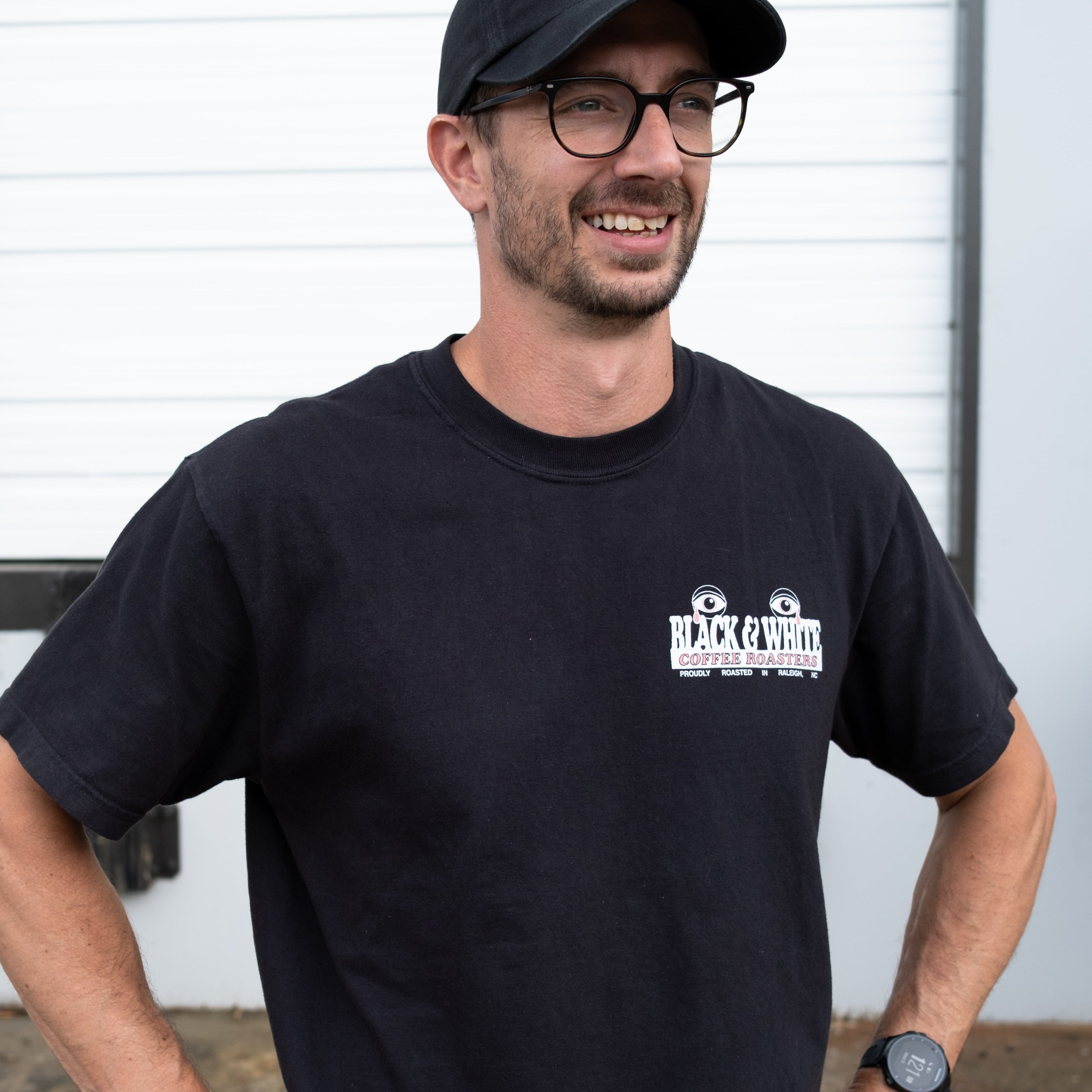 A male modeling the front of a black shirt with his hands on his hips. The front pocket of a black t-shirt showing a graphic of typography that states "Black & White Coffee Roasters, proudly roasted in Raleigh, NC." The graphic has two eyes crying red tears.