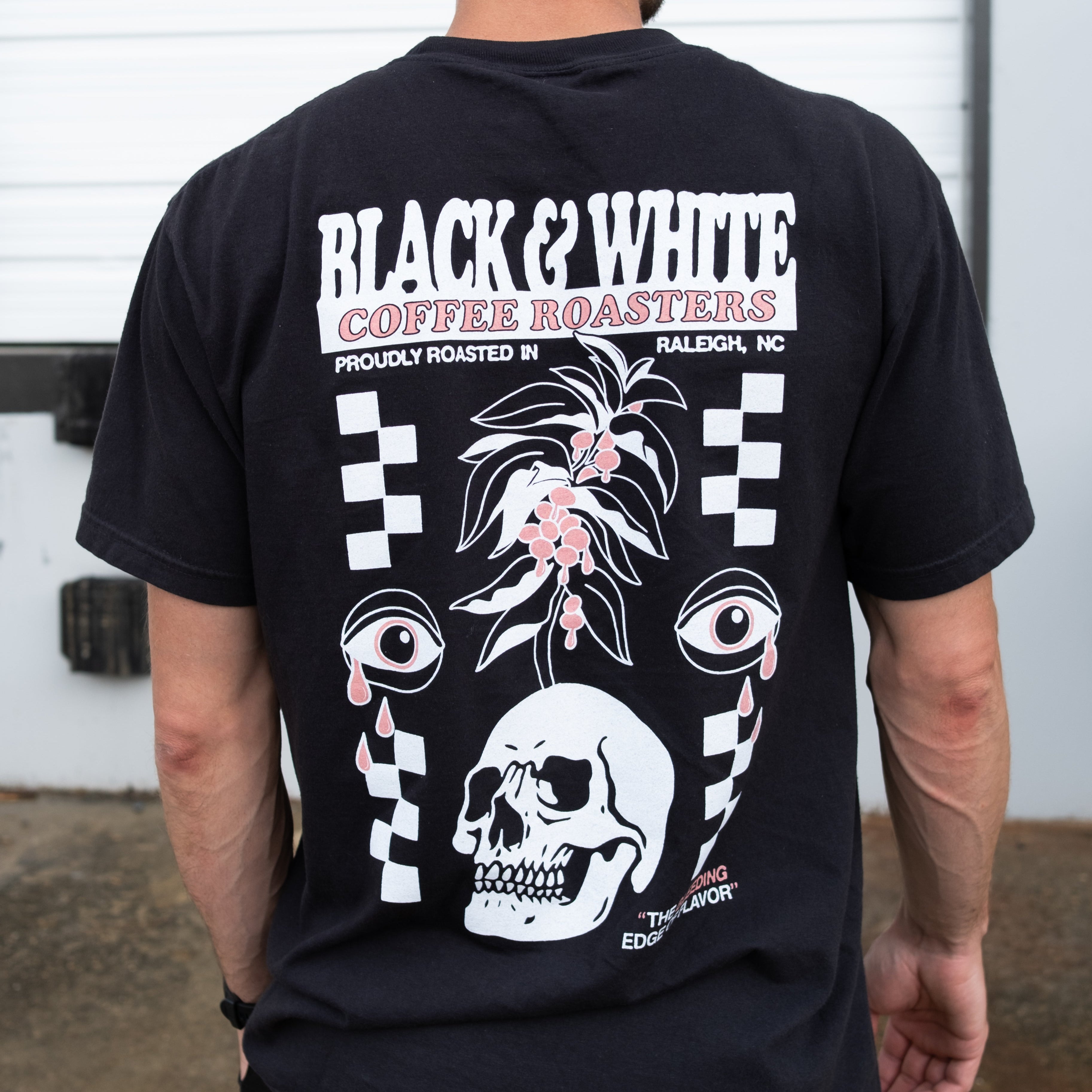 The backside of a black t-shirt being modeled on a male. The graphic is 90% of the backside of the shirt. The graphic says, "Black and White Coffee Roasters, Proudly roasted in Raleigh, NC" and "The Bleeding Edge Of Flavor." The graphic has checker board design on the left and right with a skull with a coffee tree growing out of it. The graphic is finished off two eyeballs crying red tear drops.