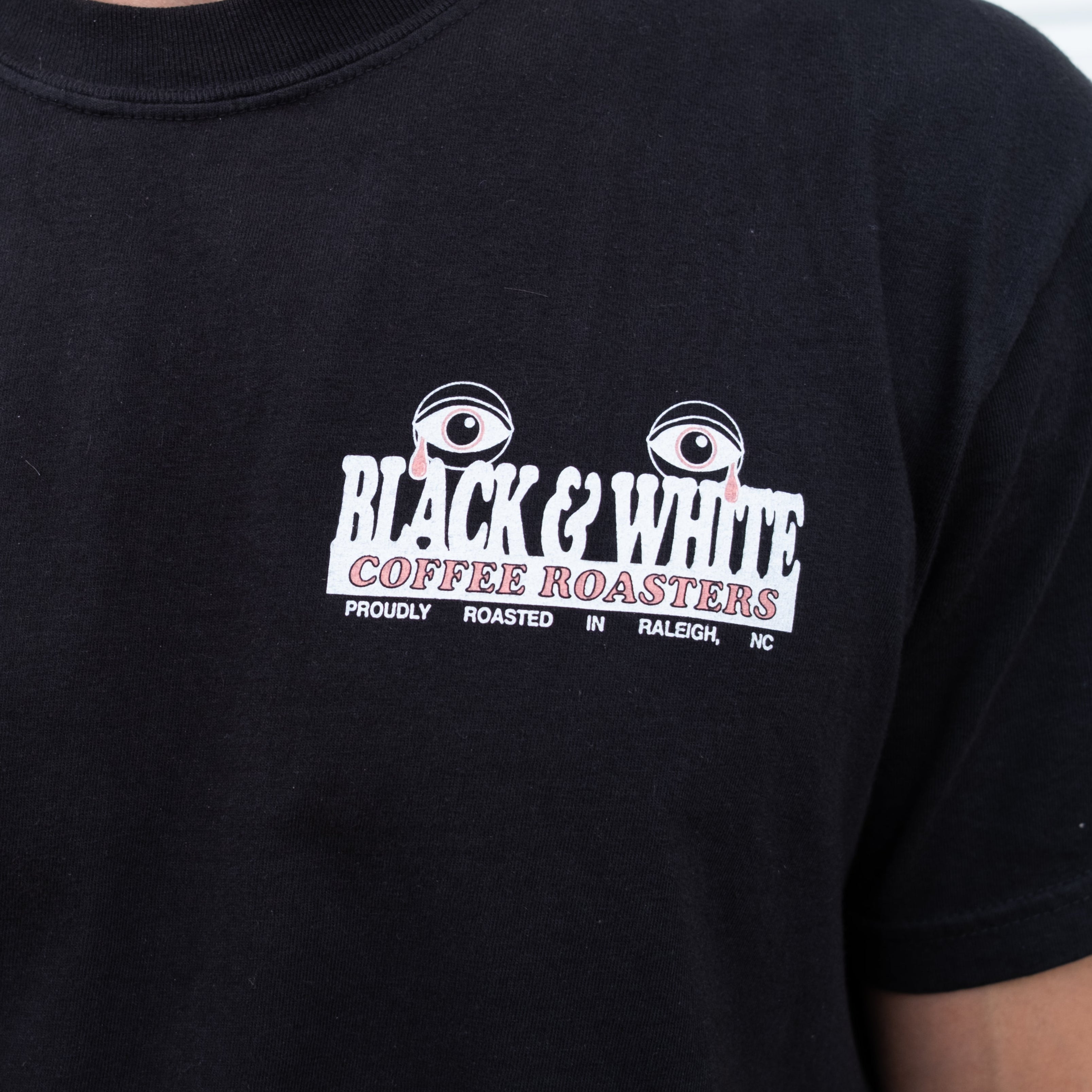 The front pocket of a black t-shirt showing a graphic of typography that states "Black & White Coffee Roasters, proudly roasted in Raleigh, NC." The graphic has two eyes crying red tears.