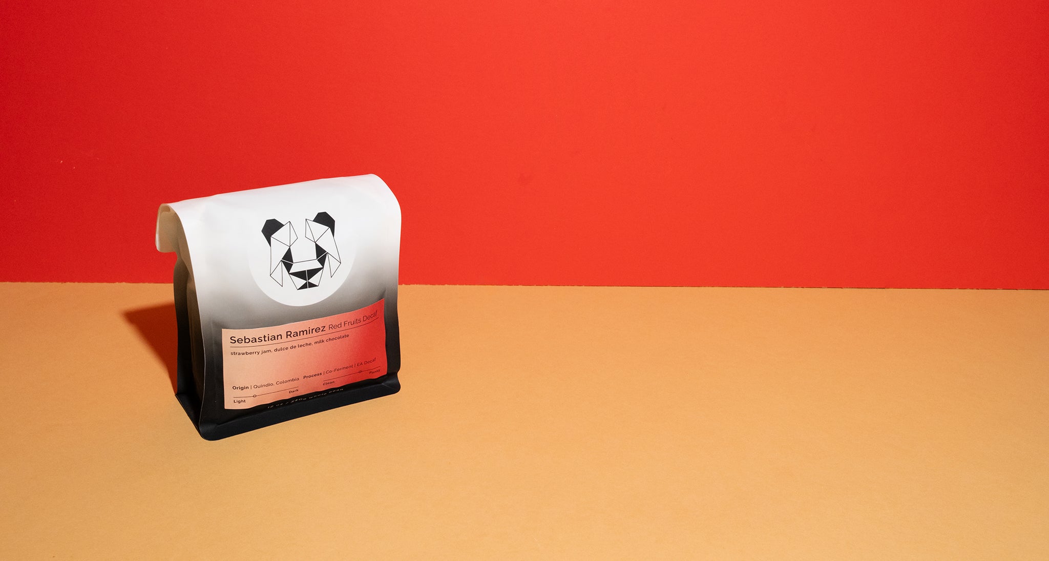 A coffee bag package with a white top flap featuring a black panda logo and a red label, standing on a beige surface against a red background. The label says Sebastian Ramirez Red Fruits Decaf.