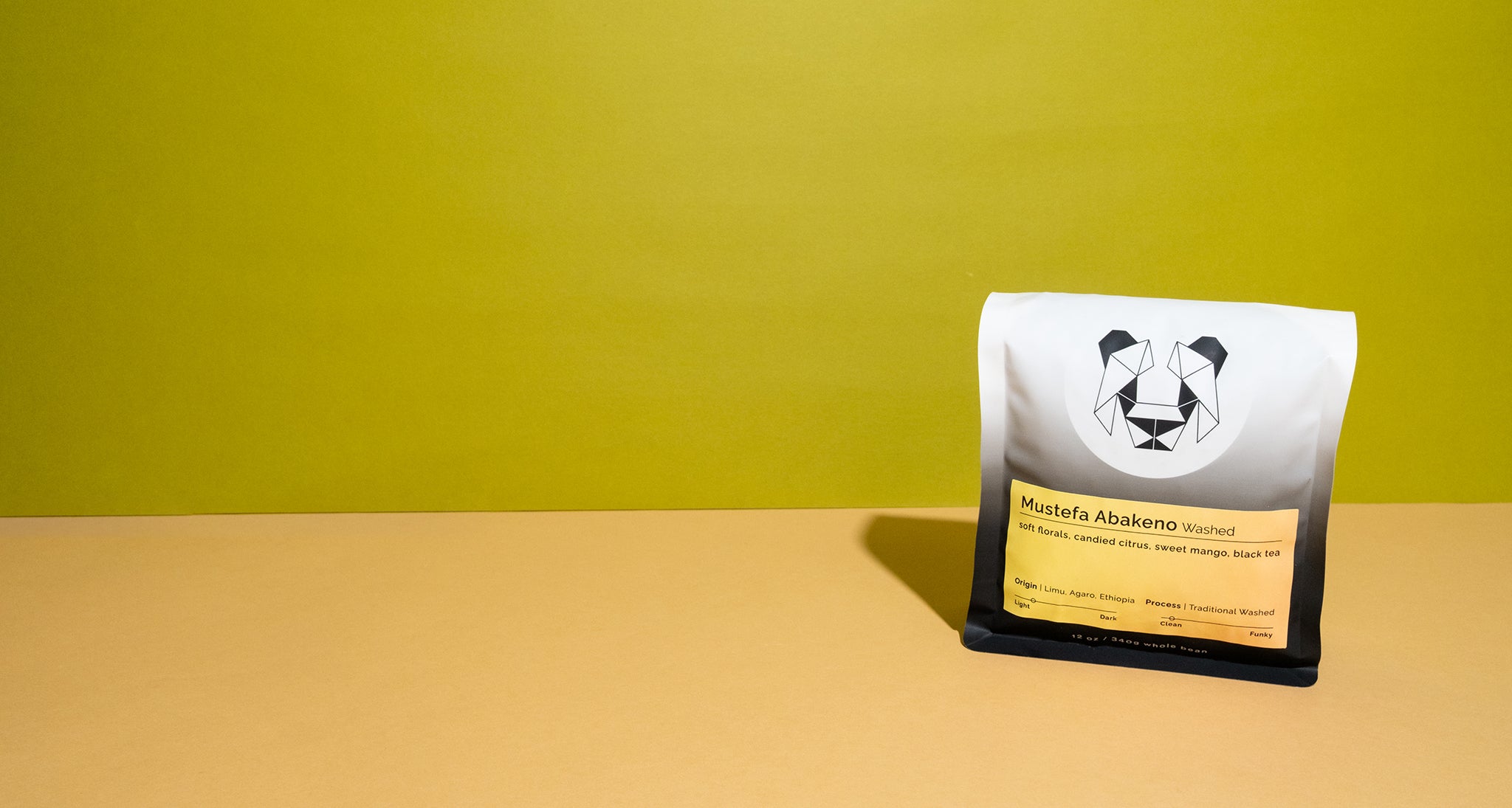 A single bag of coffee stands on a beige surface against a bright yellow-green wall. The bag is mostly white and black with a yellow label reading 'Mustafa Abakano Washed' and a panda logo.