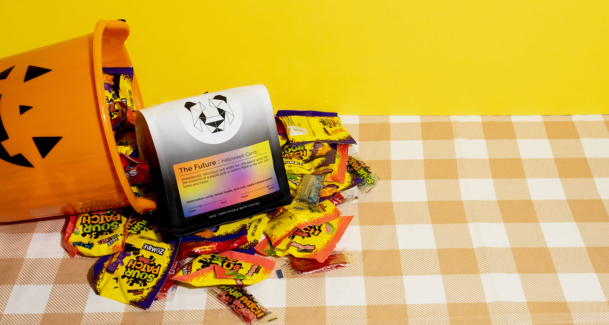 A bag of coffee with a multicolored label on a picnic table cloth with a jack o lantern candy bucket spilling out. The coffee says the future Halloween candy.