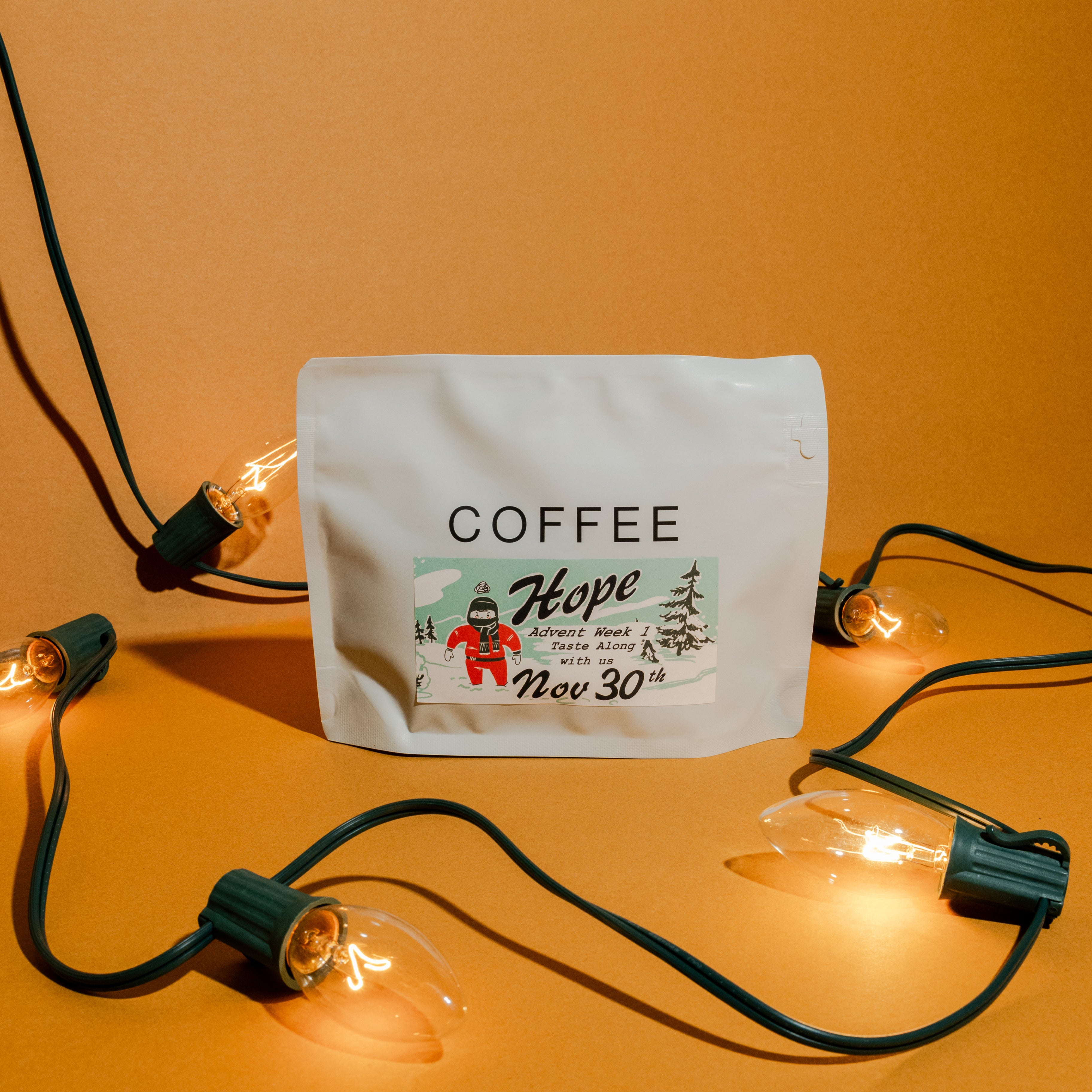2025 B&W Advent Calendar: White resealable coffee pouch with festive “Hope – Advent Week 1” sticker, surrounded by vintage-style C9 bulbs on green wire.