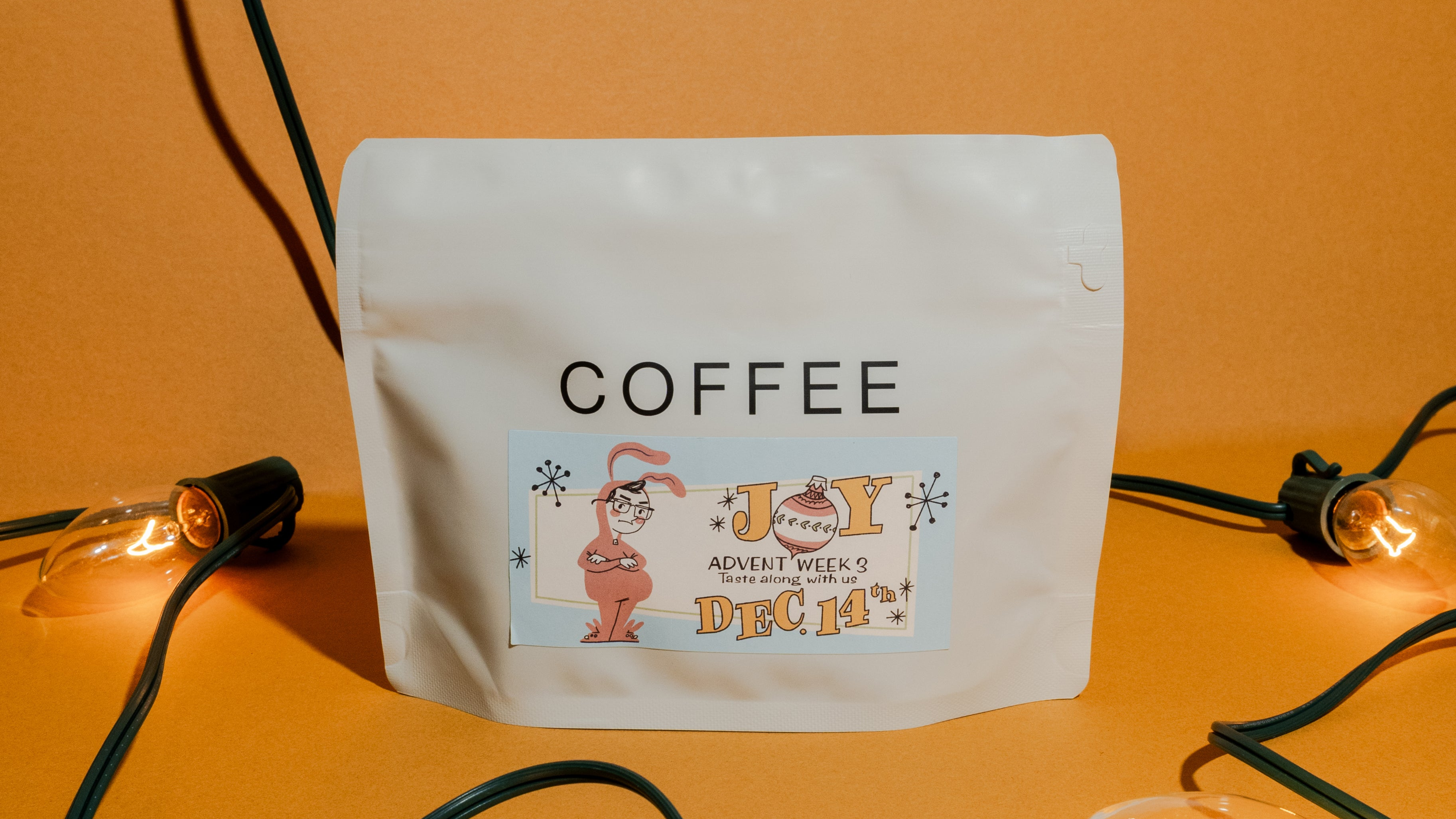 Bag of coffee on a Christmas light themed backdrop with the words Joy on the label, Advent week three. The label has a kid in a bunny costume illustrated on it.
