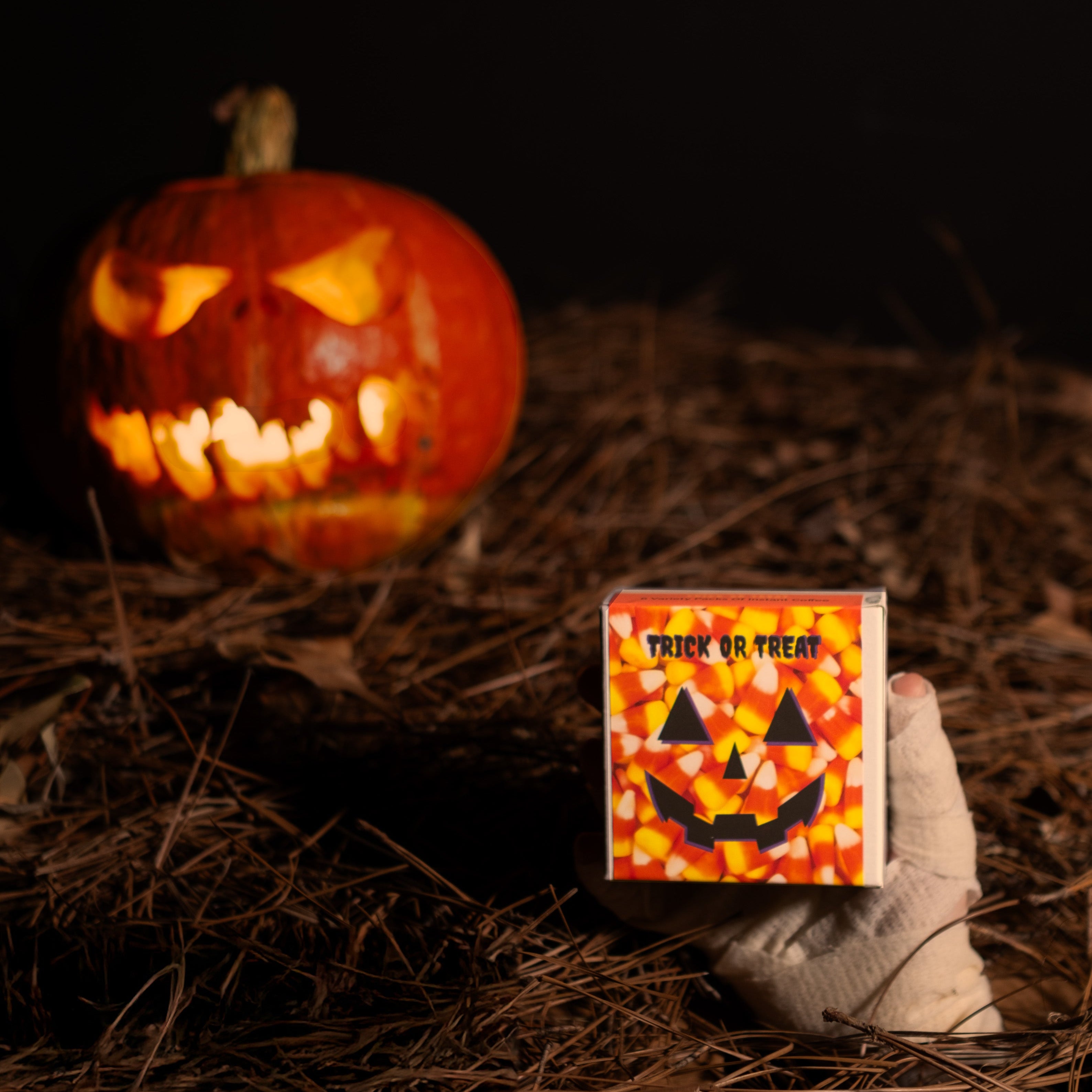Trick or Treat Variety Pack - Instant Coffee in candy-corn box, grabbed by a mummy hand, with glowing jack-o’-lantern, set in a spooky graveyard scene.